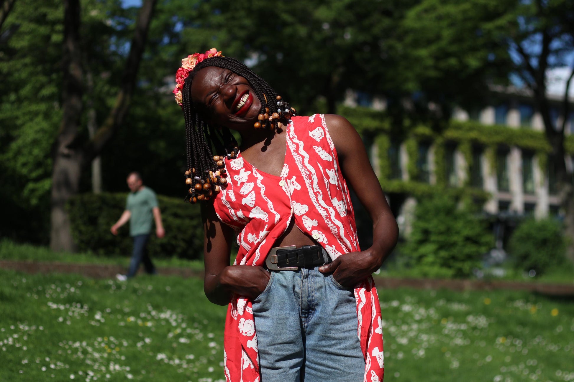 A female model with long, beaded braids smiles while wearing a red and white floral sleeveless blouse outdoors in a grassy area. Her hands are in her pocket and she shows off a floral crown in red and pink