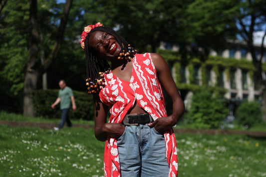 A female model with long, beaded braids smiles while wearing a red and white floral sleeveless blouse outdoors in a grassy area. Her hands are in her pocket and she shows off a floral crown in red and pink