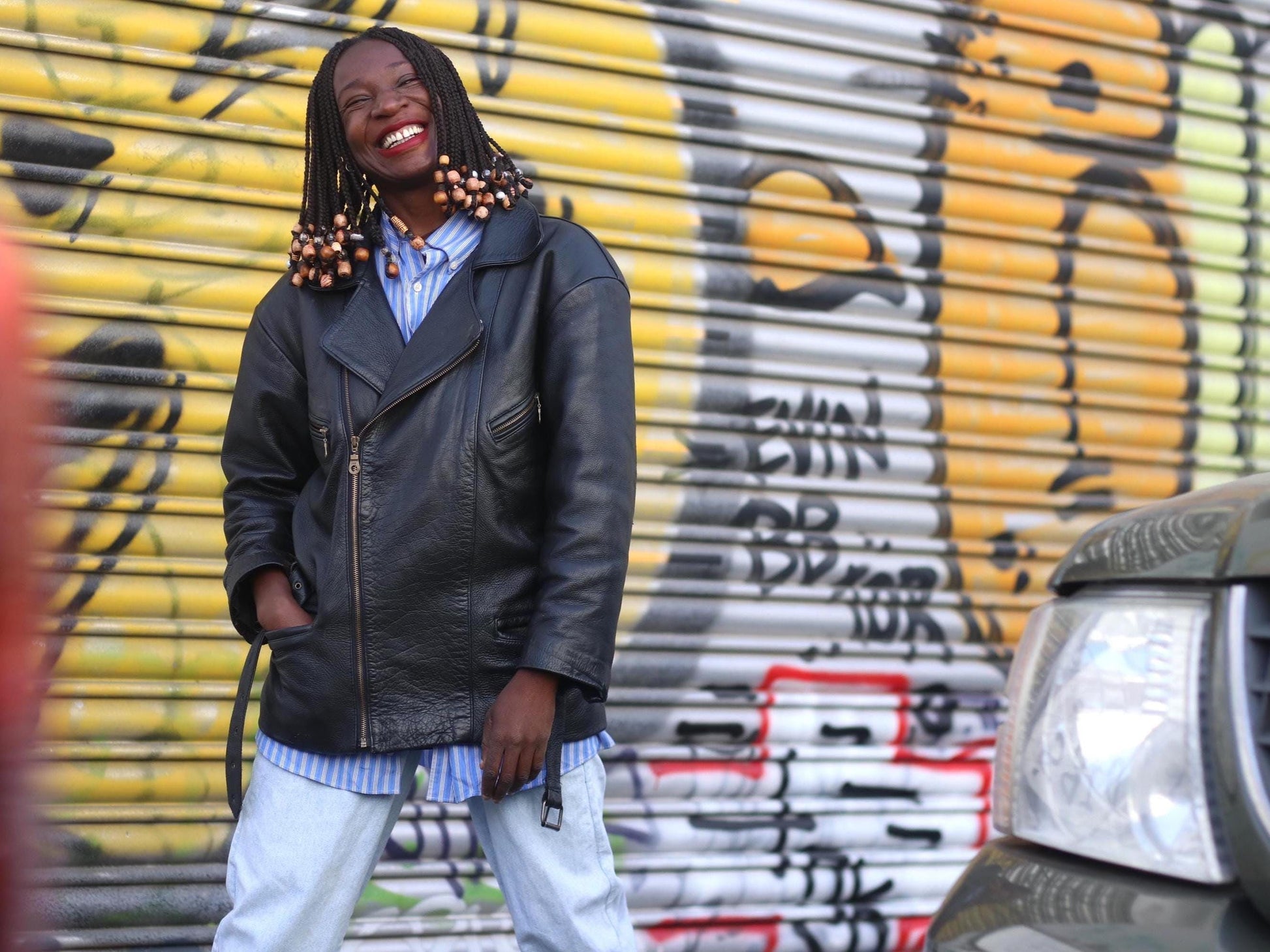Female model with braids wearing a black vintage Rock and roll style leather jacket from the 90s is photographed outdoors against a graffiti covered wall with smiling. She has a striped blue shirt underneath paired with light blue jeans.