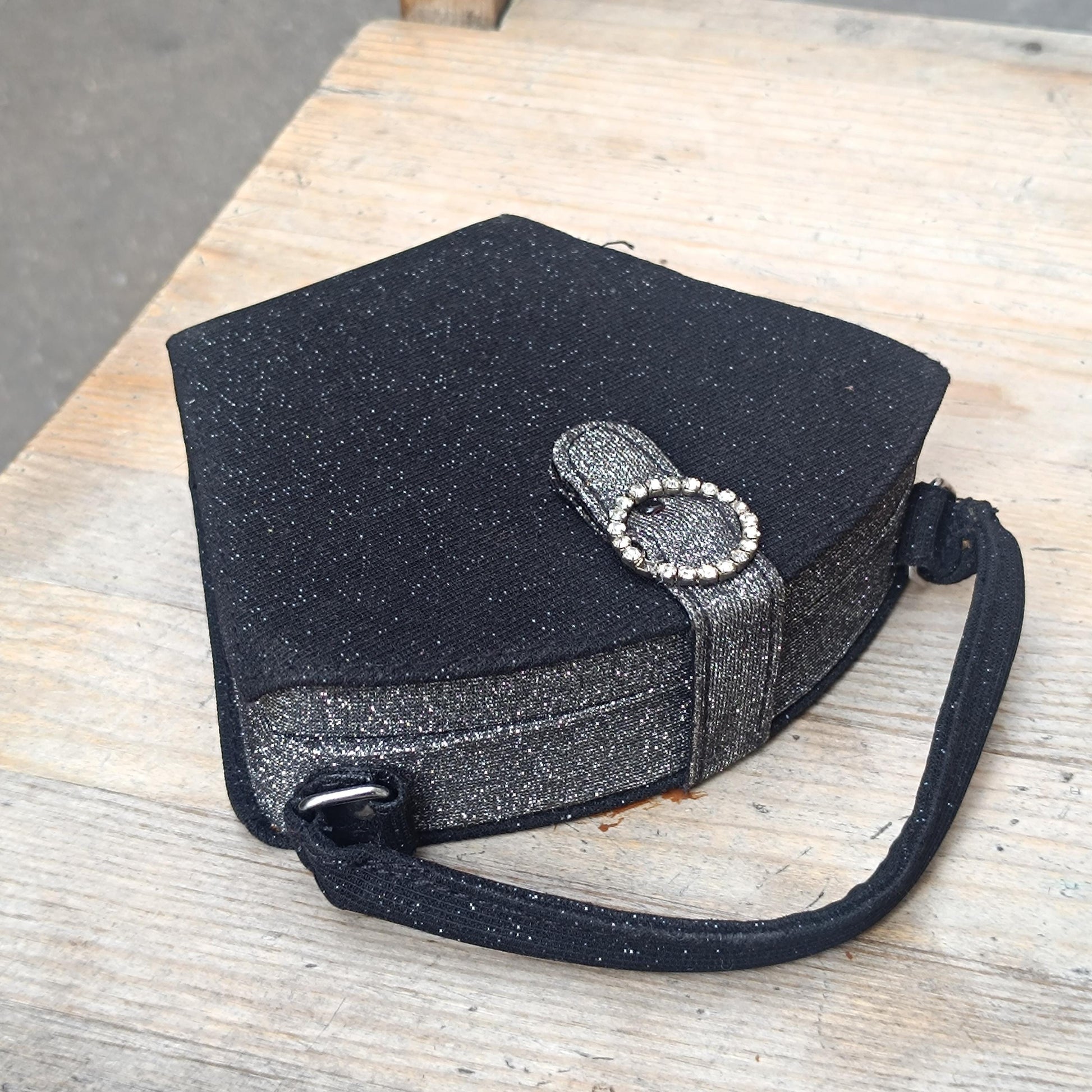 Close-up of a black glitter y2k mini bag with a circular faux-bling embellishment on the tab closure. The compact vintage bag&#39;s top handle and pentagon shape are also visible against a rustic wooden backdrop.&quot;