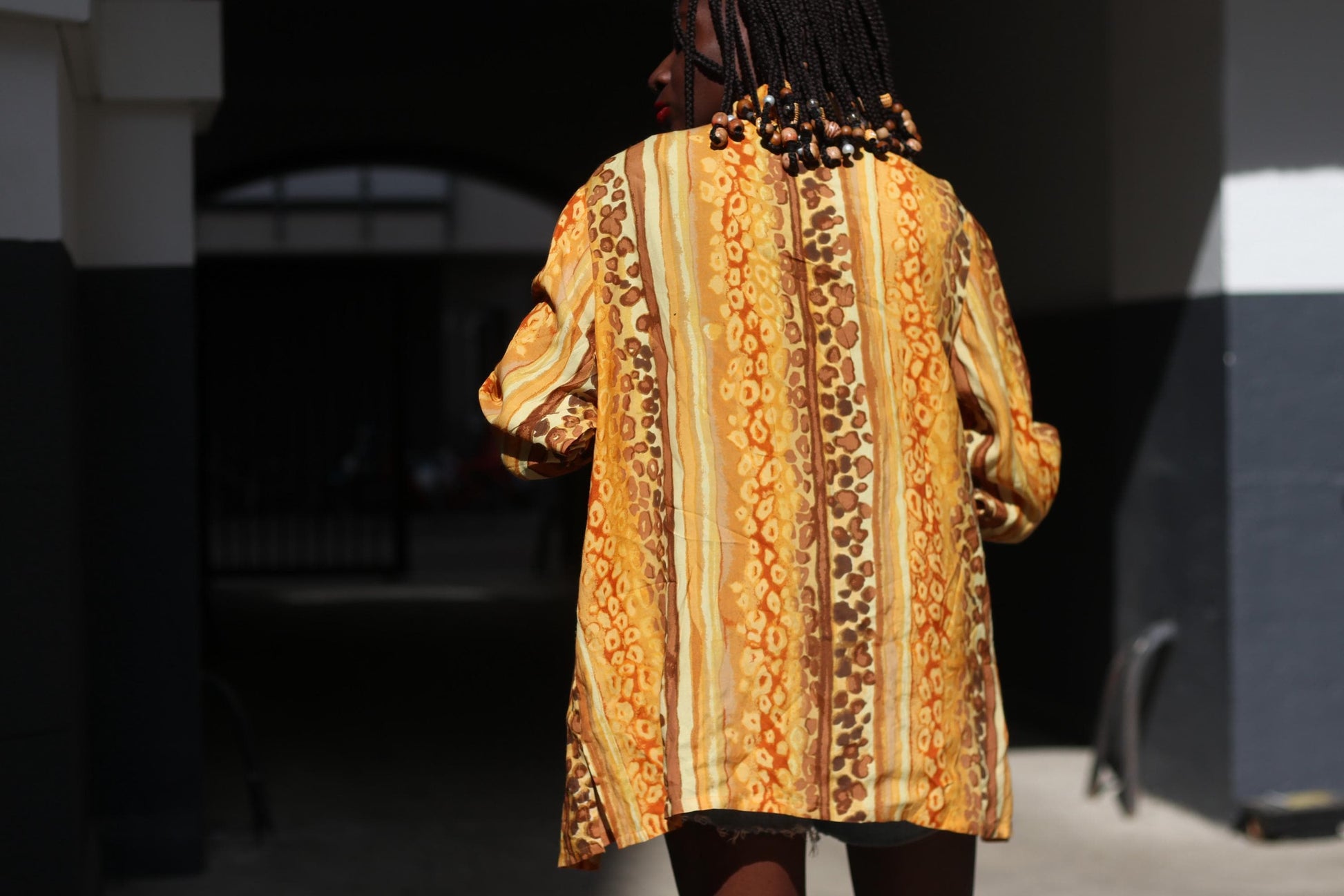 A 90s retro blouse featuring a bold animal print with swirling patterns in shades of yellow, gold, brown, and cream is modelled by a woman with braids. She is photographed outdoors with white grey walls in the background. The image is a back view of the model and the focus is on the vintage button-up. its sleeves,  and a loose, oversized fit.