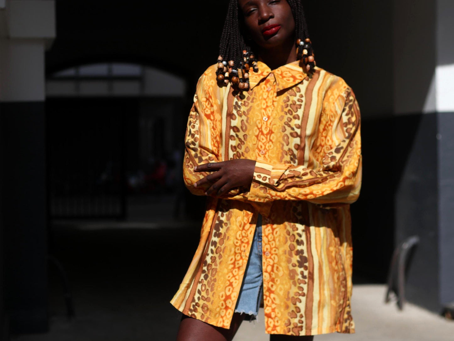 An image of a female model  wearing a yellow patterned blouse and blue jeans shot in moody lighting in an urban alleyway.The focus is on the animal-print of the 90s blouse and the oversized silhouette