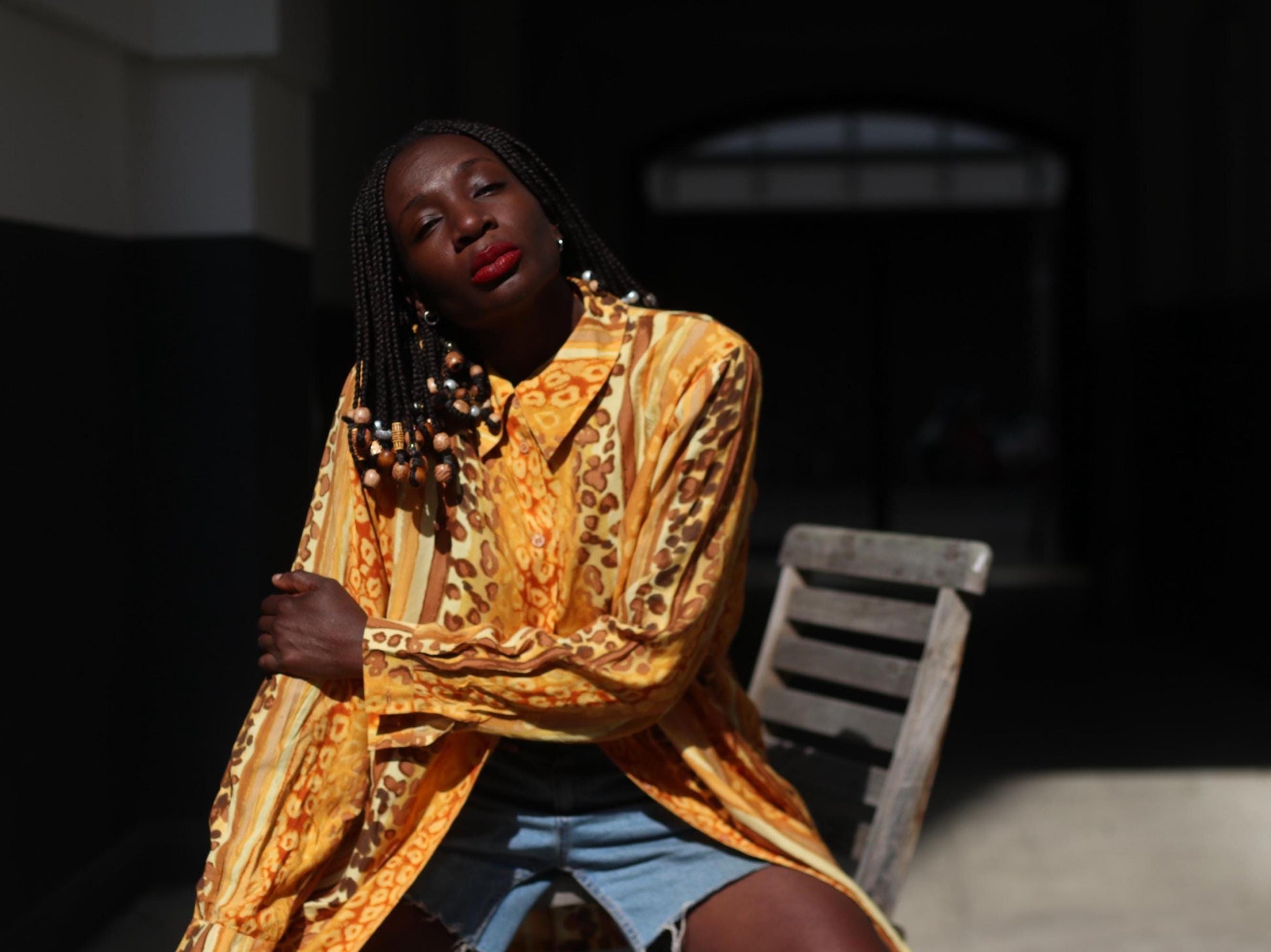 An Outdoor shot of a female model wearing a yellow patterned blouse and blue denim shorts. The.woman sits on the arms of a light grey chair  modeling the vintage oversized 90s blouse. She has one hand on the elbow of the other hand and looks directly into the camera. The outline of a gate is further in the background