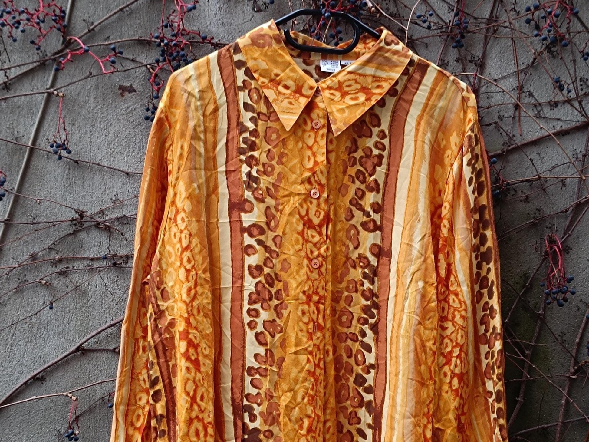 A vibrant 90s yellow animal print blouse with swirling patterns in shades of yellow, gold, brown, and cream is photographed hanging outdoors against a weathered grey  wall with brown twines. The blouse has long sleeves, a button-down front, and a loose, oversized fit.