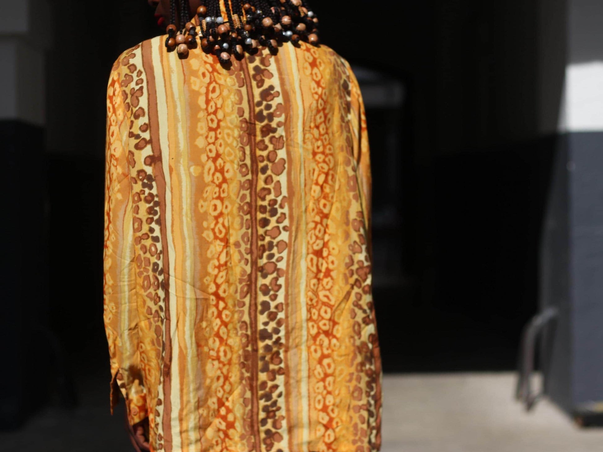 The back of a  90s blouse with a bold animal print with swirling patterns in shades of yellow, gold, brown, and cream modelled by a woman of color with braided beads. The blousehas long sleeves and a loose, oversized fit.