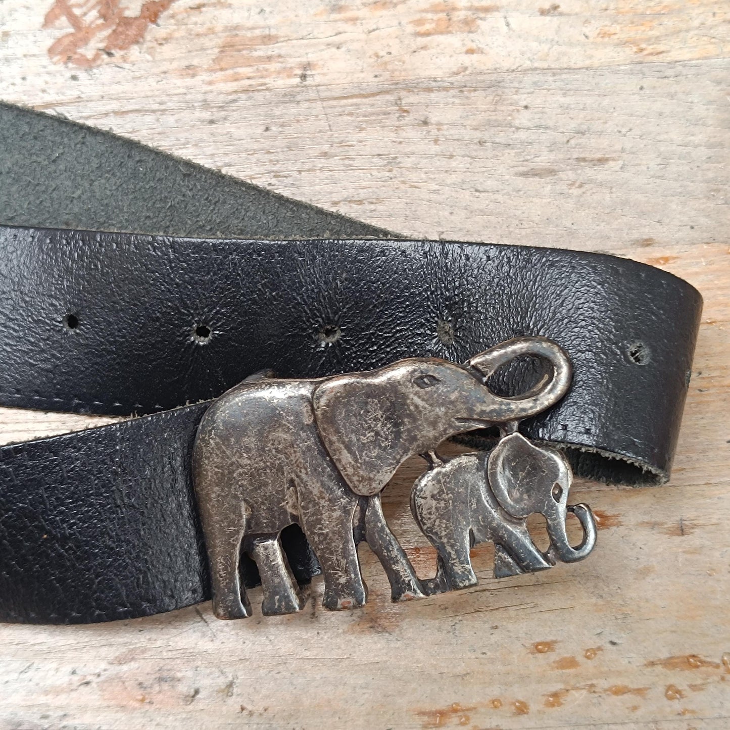 Close-up of a vintage black leather belt with an antique silver elephant buckle, showcasing intricate front detailing. The 70s handmade piece is photographed on a  wooden brown rustic surface