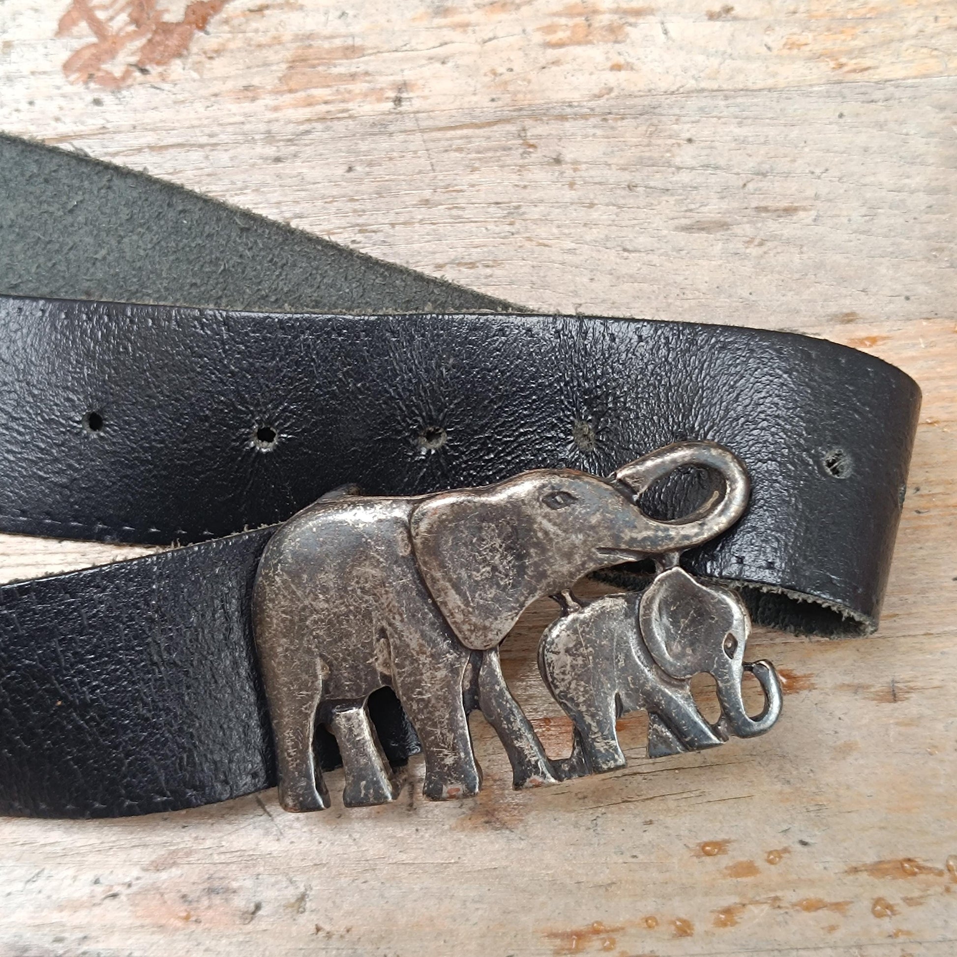 Close-up of a vintage black leather belt with an antique silver elephant buckle, showcasing intricate front detailing. The 70s handmade piece is photographed on a  wooden brown rustic surface