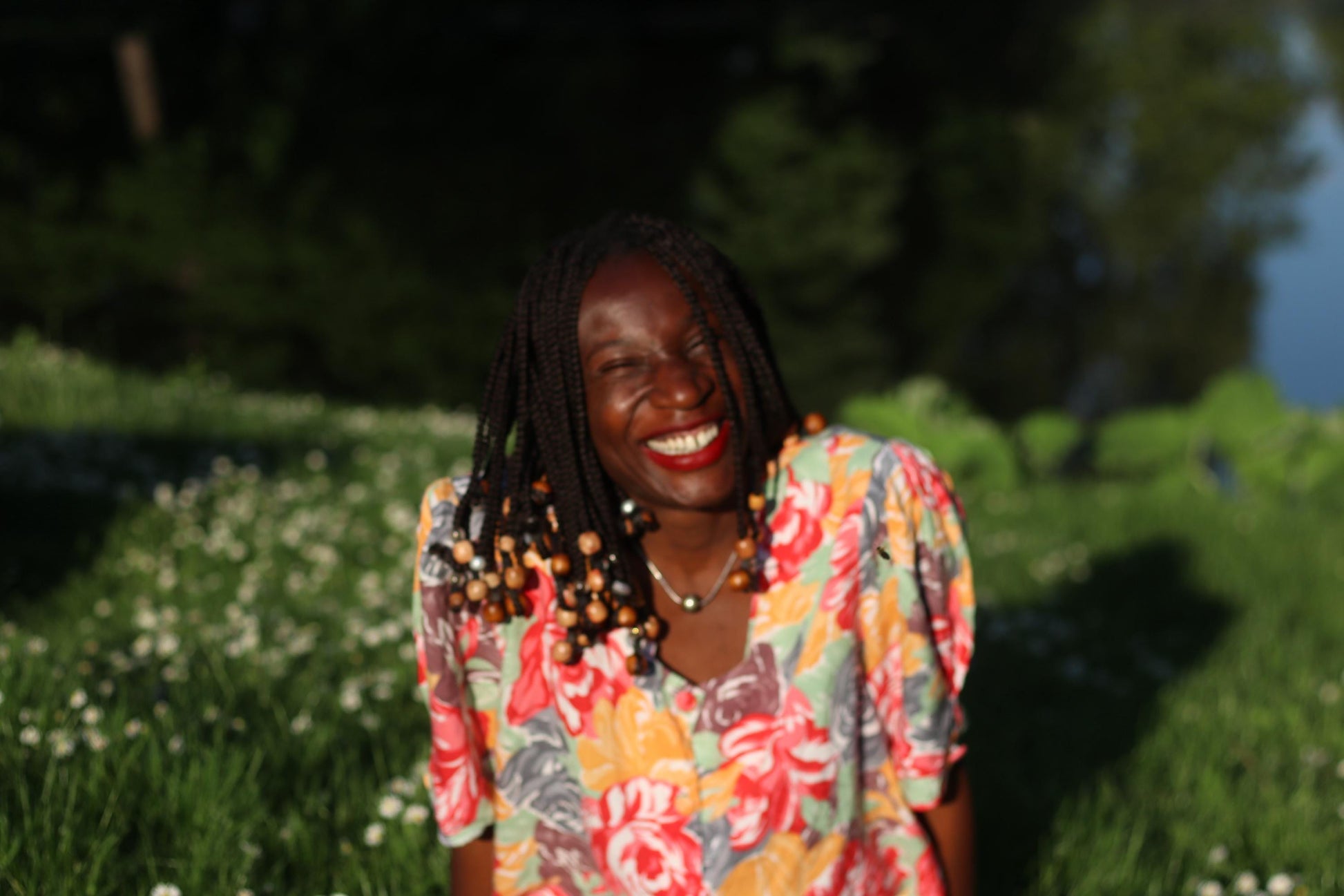 Smiling model with beaded braids wearing a vintage floral blouse with puff sleeves is photographed sitting in the grass, looking into the camera on a sunny day. Background is  compromised of greenery and scattered white daisies