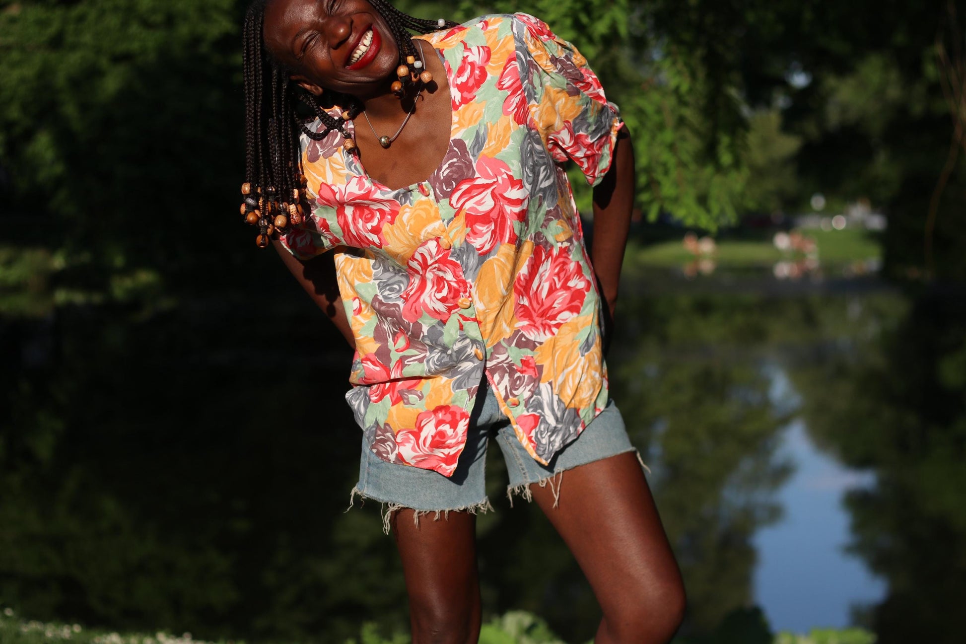 Smiling model with beaded braids wearing a vintage retro floral blouse with puff sleeves paired with denim shorts  is photographed outdoors Infront of a body of water. The blouse is a retro piece by H&M