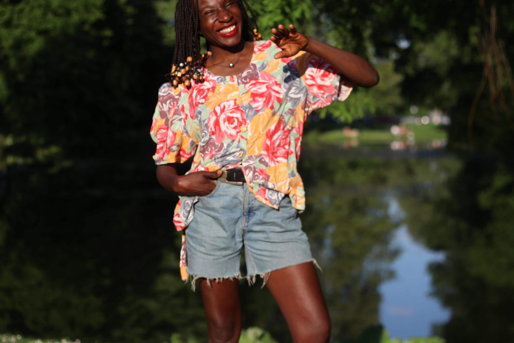 Smling model with beaded braids wearing a vintage floral blouse with puff sleeves combined with blue denim shorts is photographed sitting in the grass, looking into the camera on a sunny day. Background is greenery