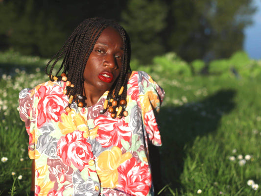Model with beaded braids wearing a vintage floral blouse with puff sleeves is photographed sitting in the grass, looking into the camera on a sunny day. Background is greenery