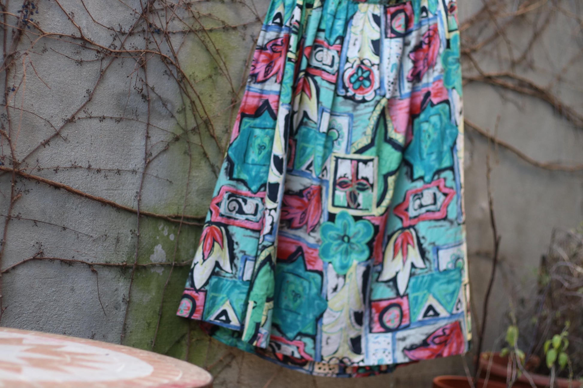 A still life image of a brightly patterned 80s vintage midi skirt is displayed on a hanger against a rustic stone wall with overgrown vines
