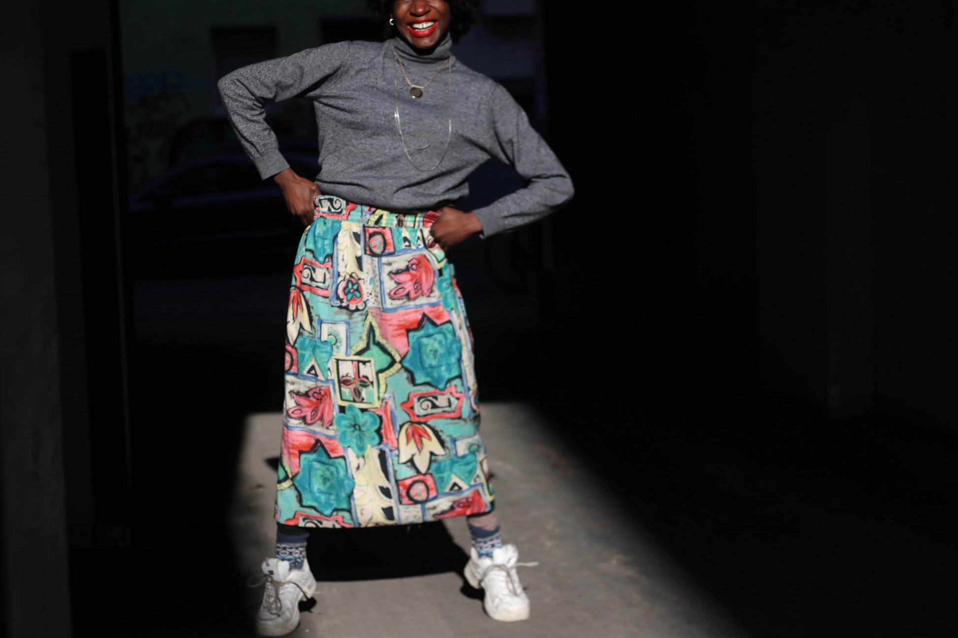 A street style photo of a female model with curly black hair wearing  a long,colorful  vintage skirt with a unique, artistic print against a dark, shadowy background. They are dressed in a gray sweater and white sneakers hands in their hips
