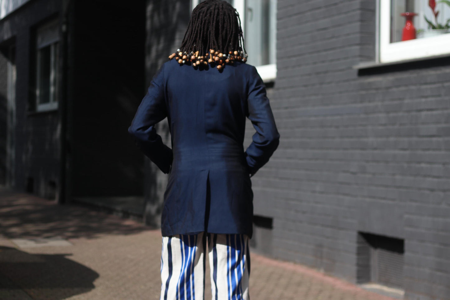 A  back view of a female model with beaded braids photographed on a sunny day against a brick wall on a sunny day in a vintage 90s blue blazer by the brand Bonita styled with blue and white striped cullotes . The look is a vintage twist on power dressing.
