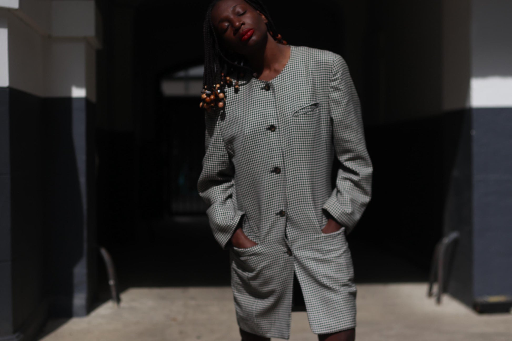 Front view of a vintage black and white checkered blazer dress with structured shoulders and oversized buttons, styled as a statement outerwear piece on a female model with beaded black braids in an urban outdoor environment. model has both hands in her pocket