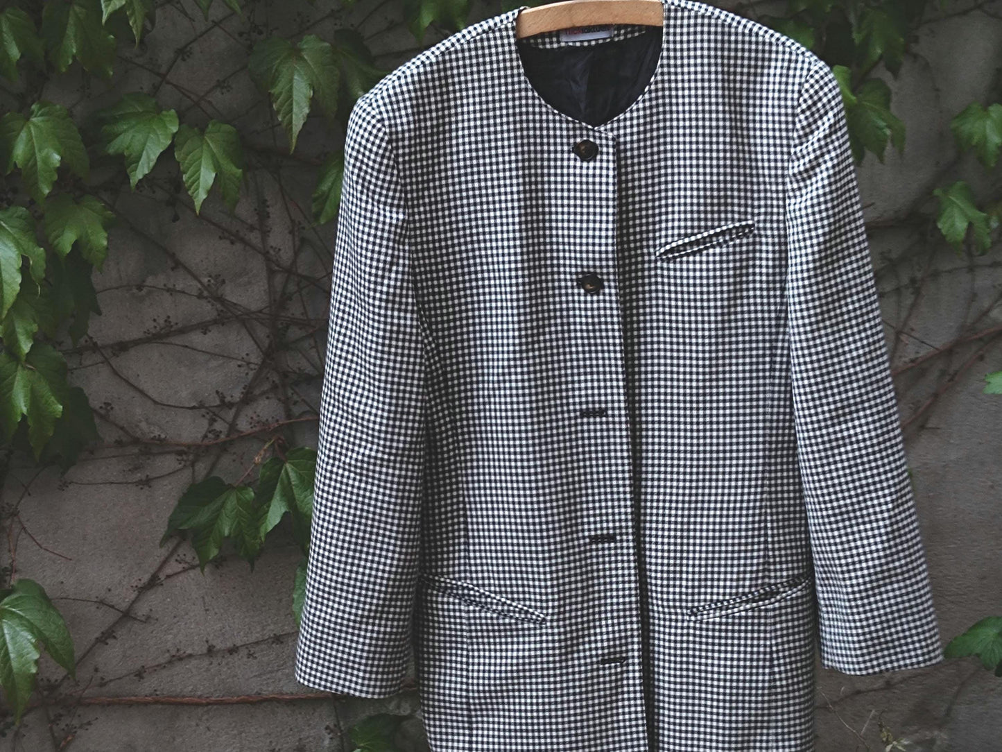 vintage black and white checkered blazer dress with structured shoulders and oversized buttons photographed outdoors on a hanger against a backdrop of a weathered grey wall with climbing plants