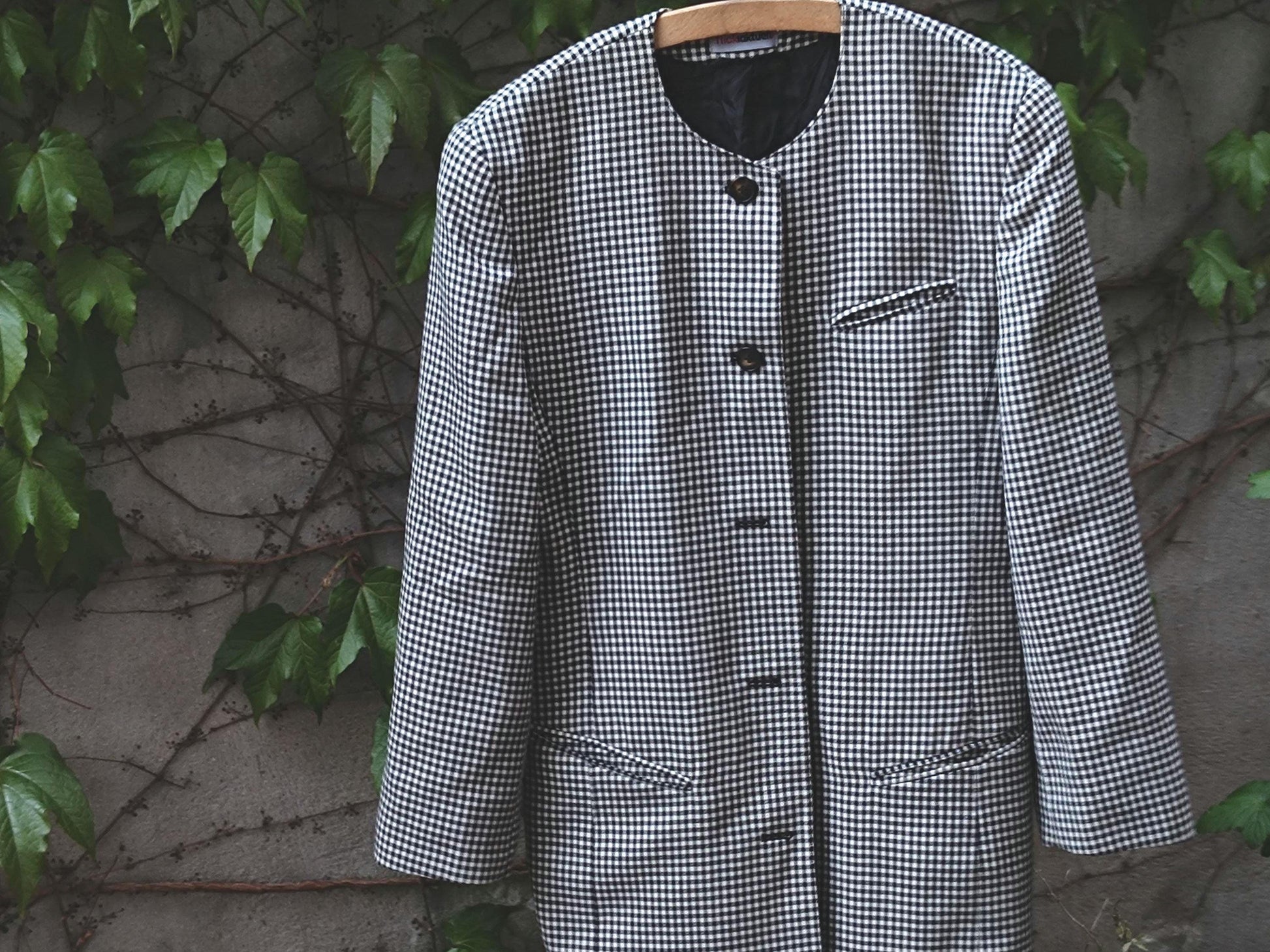 vintage black and white checkered blazer dress with structured shoulders and oversized buttons photographed outdoors on a hanger against a backdrop of a weathered grey wall with climbing plants