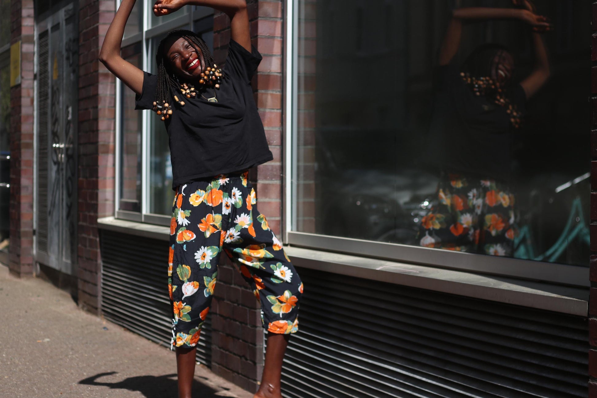 A smiling female model stands in bright sunlight wearing vintage black capris from the 90s with bold orange and white floral prints, paired with a simple black t-shirt against a brick wall and glass storefront. Her hands are raised above her head