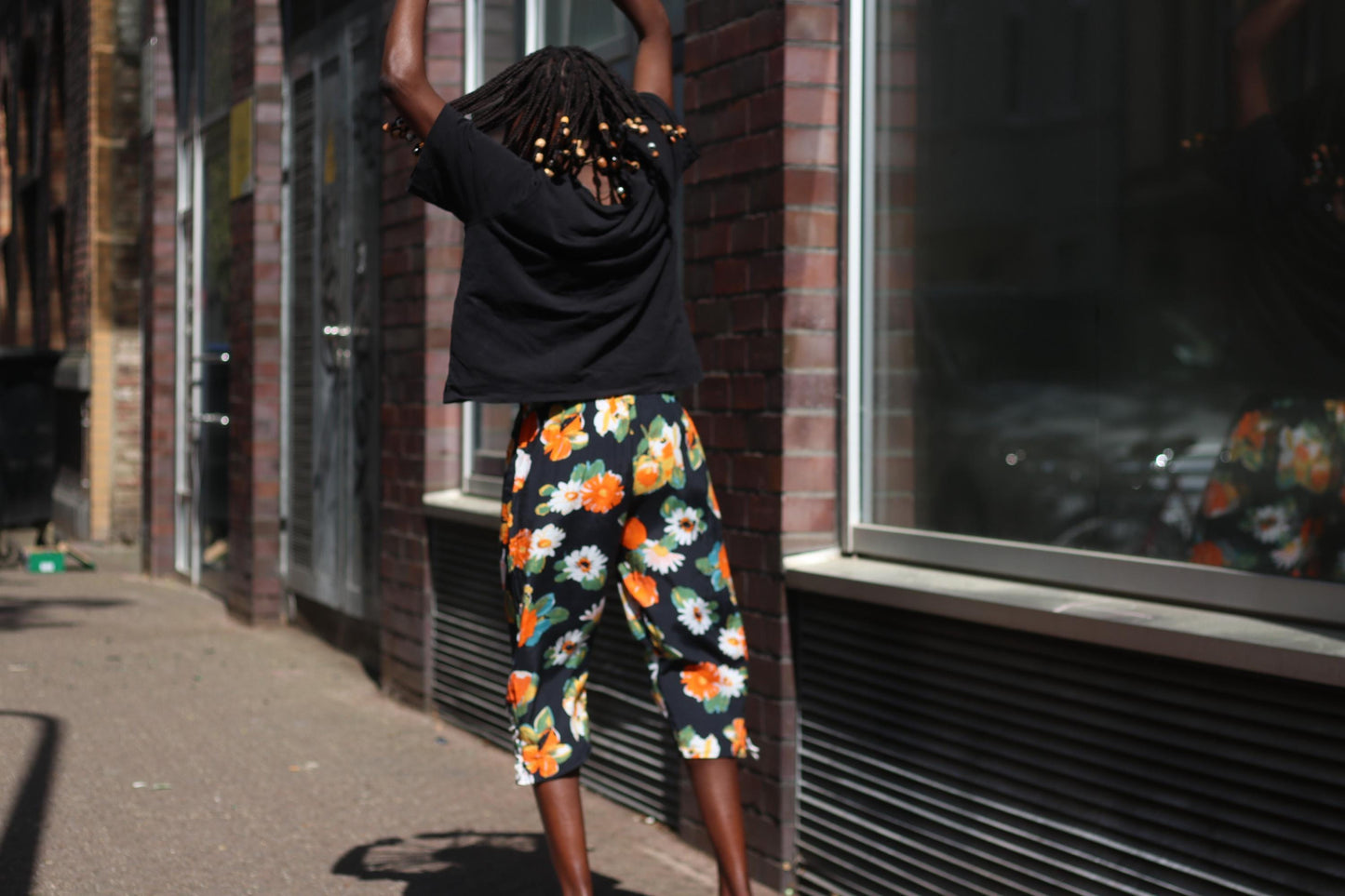 A back view of Bold floral vintage capri pants worn casually with a loose black tee, on a model of color with braided beads. Model has her hands joyfully up in the air.