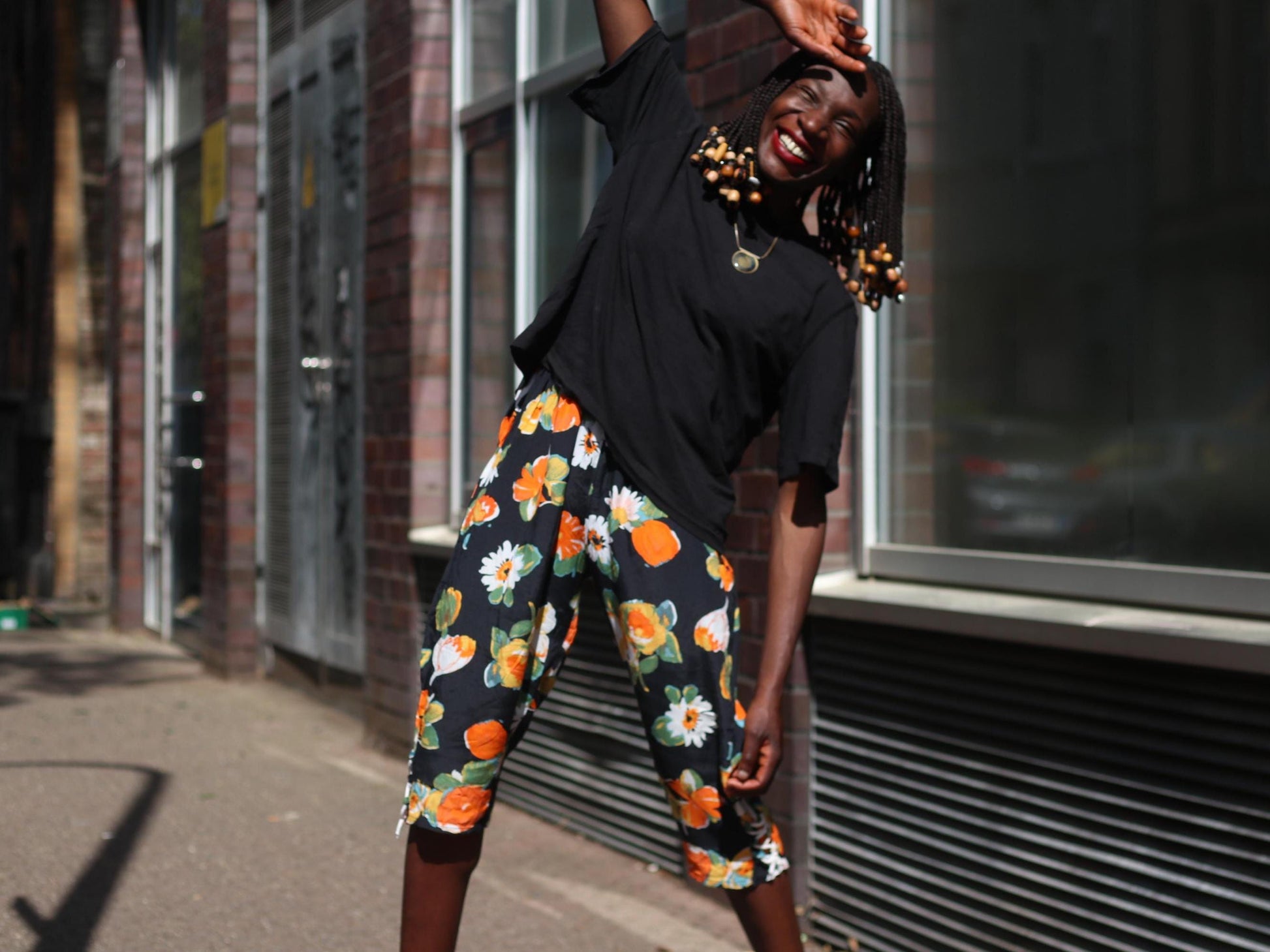 A female model stands in bright sunlight wearing vintage black capris from the 90s with bold orange and white floral prints, paired with a simple black t-shirt  against a brick wall and glass storefront.