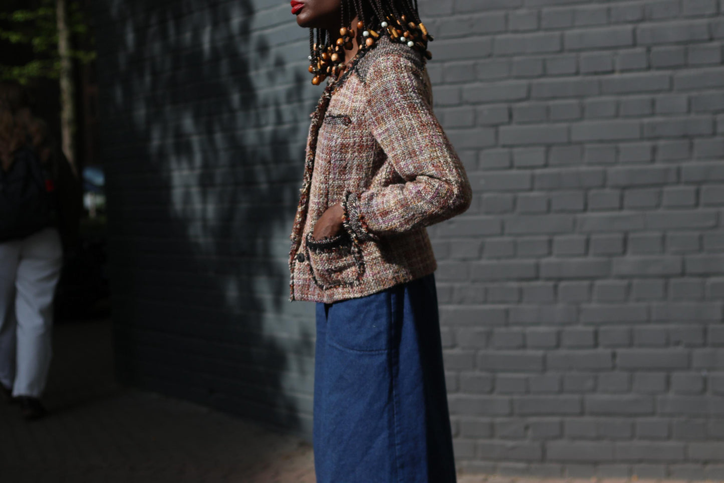 Side view Portrait of a person wearing a distinctive 90s brown tweed jacket paired with wide blue pants, set against a stark dark grey brick wall.