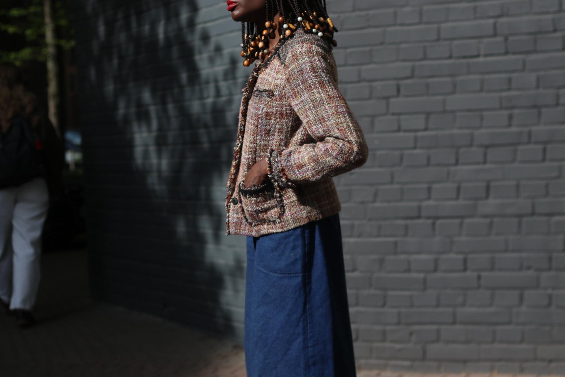 Side view Portrait of a person wearing a distinctive 90s brown tweed jacket paired with wide blue pants, set against a stark dark grey brick wall.