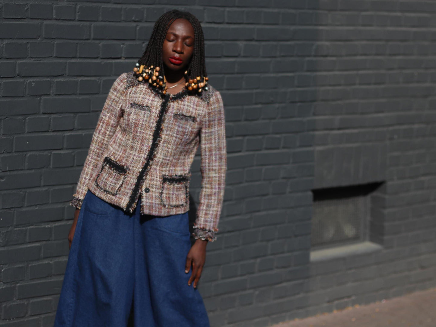 Female model with beaded braids showcasing a brown, cream, and black tweed blazer featuring a round neckline, complemented by flowing blue trousers in an urban setting.