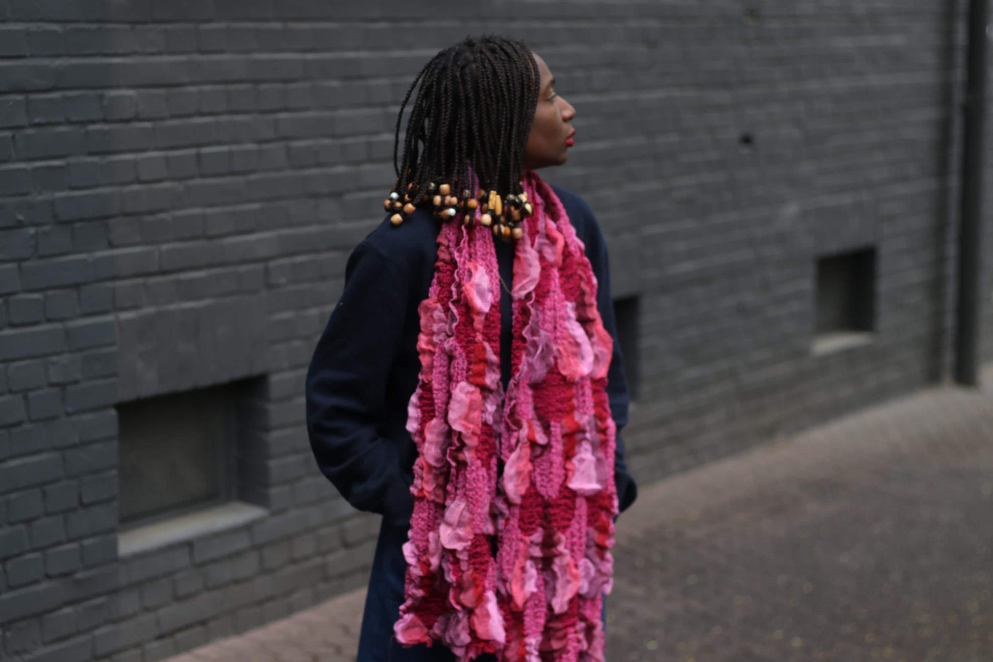 A vintage boho-style scarf in shades of pink, with a distinct knit and feel, worn by a female model against a backdrop of dark brick wall.