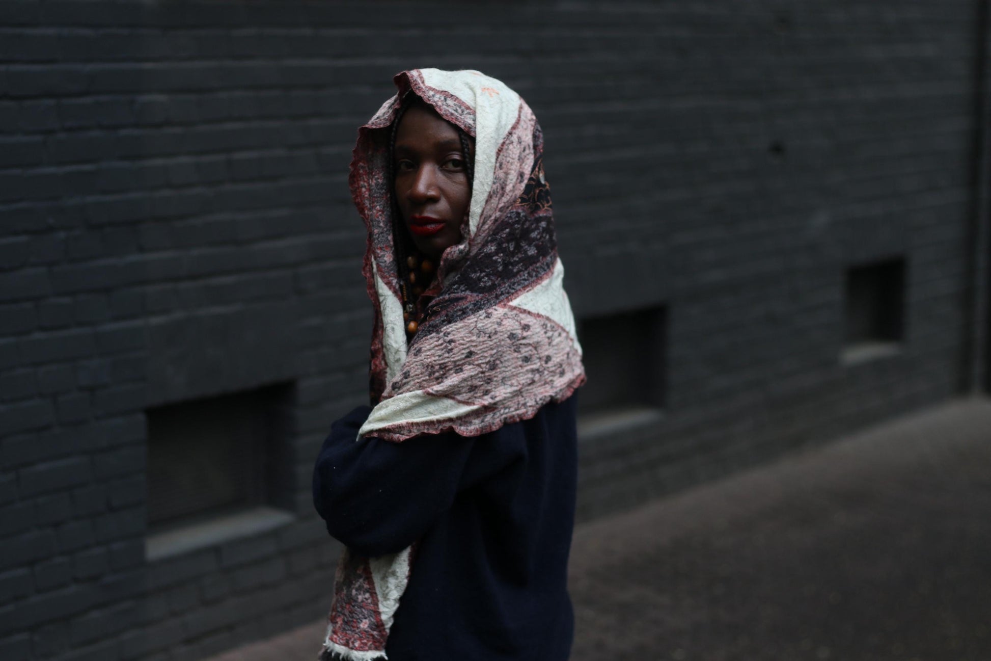 A vintage scarf from the 90s with a distinctive print worn by a female model with beaded braids as a headscarf. The model is standing in front of a dark brick wall.
