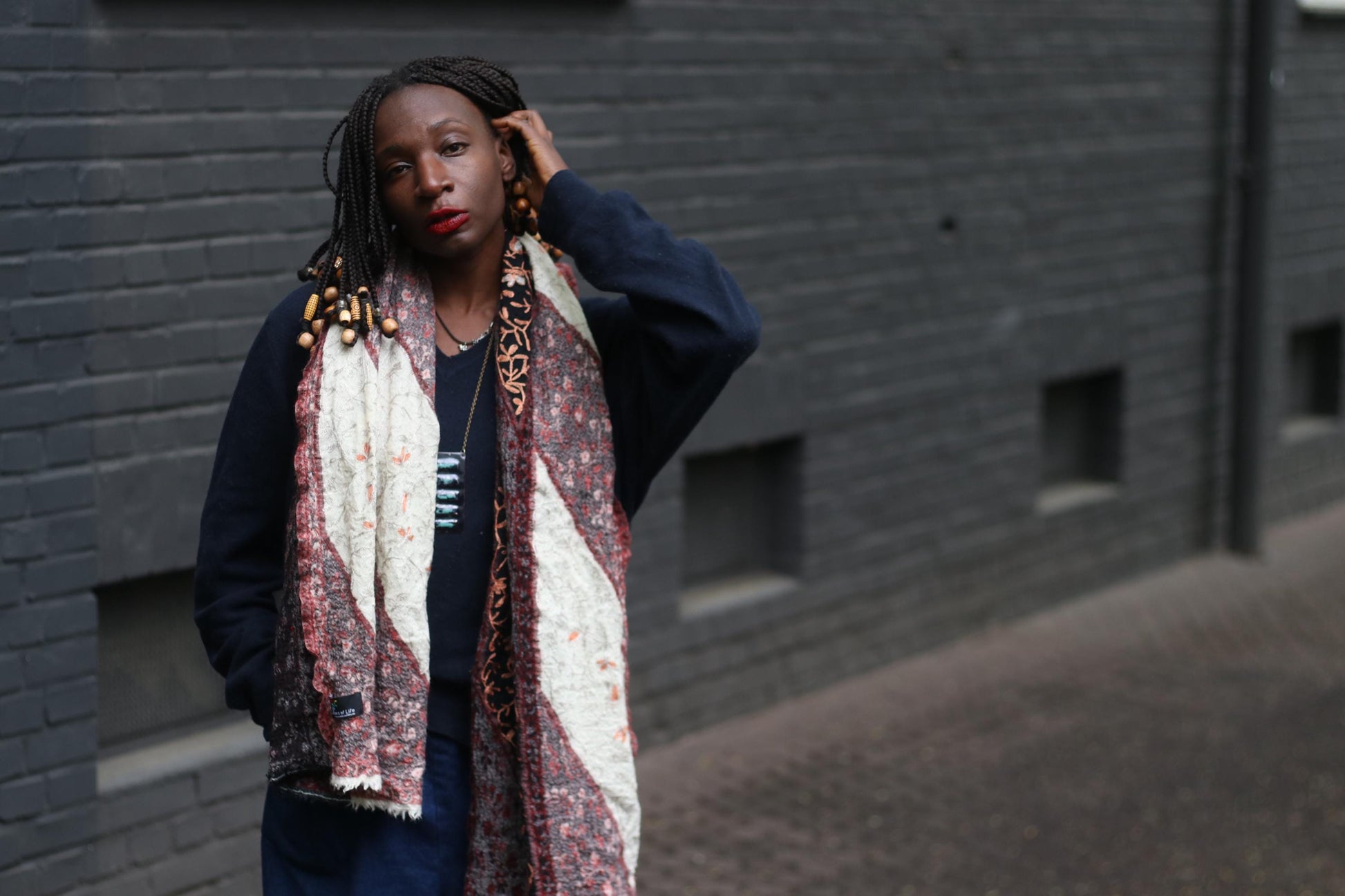Front view of a retro patterned scarf worn by a female model, the outdoor image, with a textured brick wall in the background, showcases the unique print and color palette of the boho oversized scarf.
