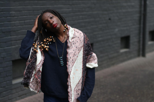 A retro oversized vintage shawl with patchwork silhouette with a distinctive print is showcased by a female model with beaded braids, standing in front of a dark brick wall. She also wears a navy sweater and a statement blue pendant necklace .