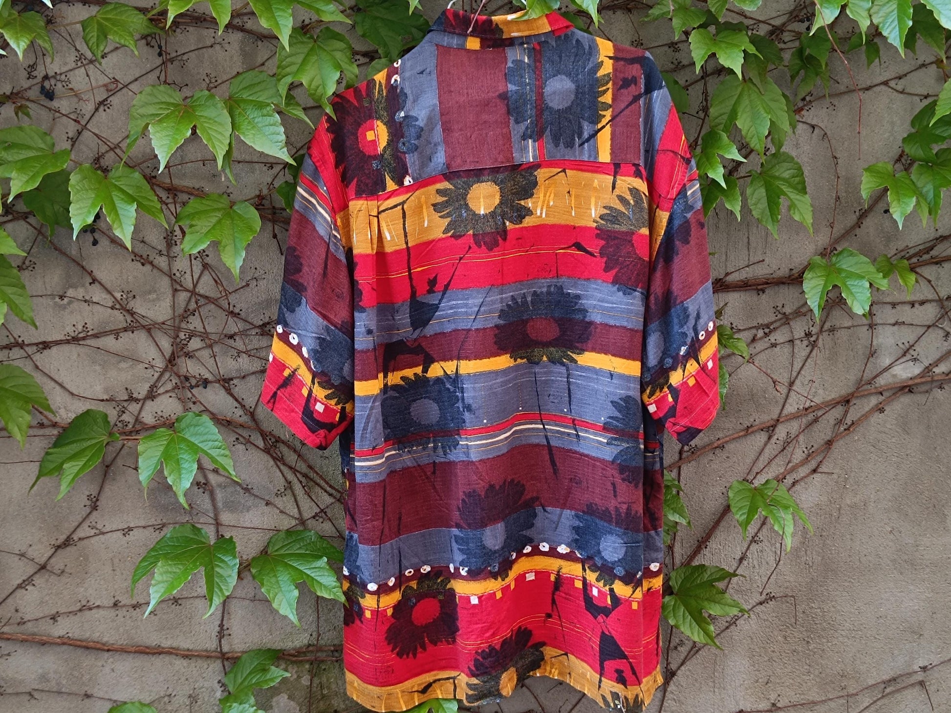 Back view of a vintage Montino tropical in rfloral shirt with bold red and yellow island print photgraphed on a hanger outdoors against a wall with climbing green plants and brown vines .