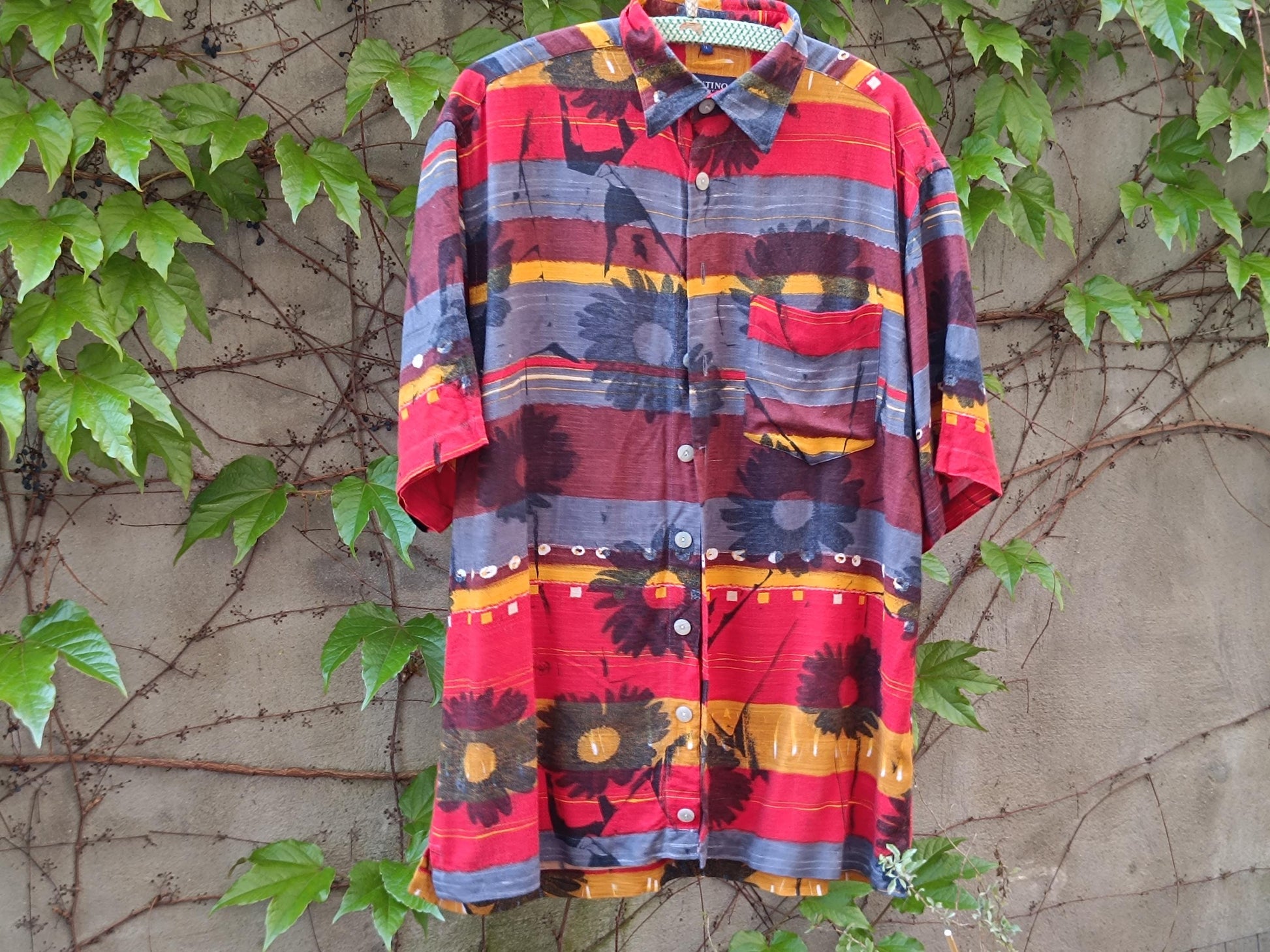 Vintage Montino tropical floral shirt with a bold island print in red and yellow photgraphed outdoors on a hanger against a wall with climbing green plants and brown vines.