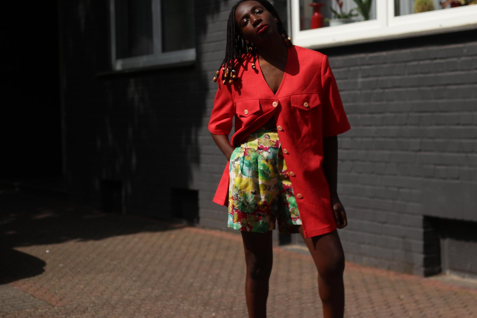 Female model wearing a vintage 90s Short-Sleeve Blazer by Mark Wald with a multicolored  pair of shorts, photographed in front of a grey brick wall.  The image showcases the  bold V-Neck Jacket as a lightweight choice for suiting up in the warmer months