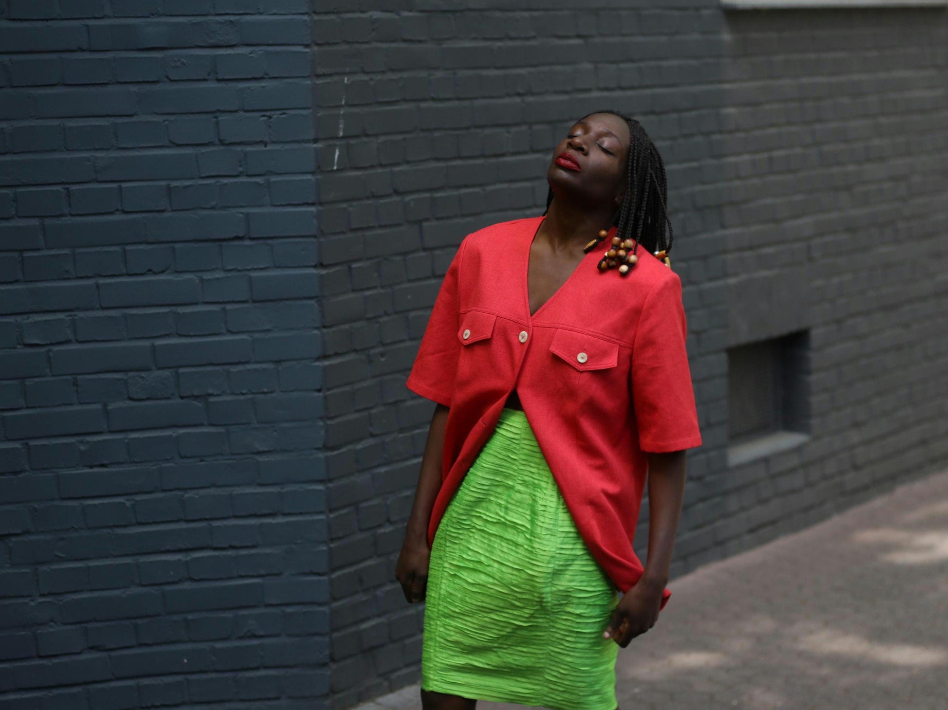 Female model wearing a vintage 90s Short-Sleeve Blazer by Mark Wald with a neon green knee-length skirt photographed in front of a grey brick wall.  The image showcases the  bold V-Neck Jacket as a lightweight choice for suiting up in the summer
