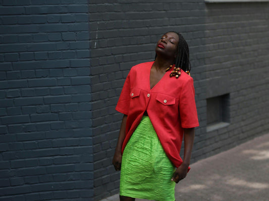 Female model wearing a vintage 90s Short-Sleeve Blazer by Mark Wald with a neon green knee-length skirt photographed in front of a grey brick wall.  The image showcases the  bold V-Neck Jacket as a lightweight choice for suiting up in the summer
