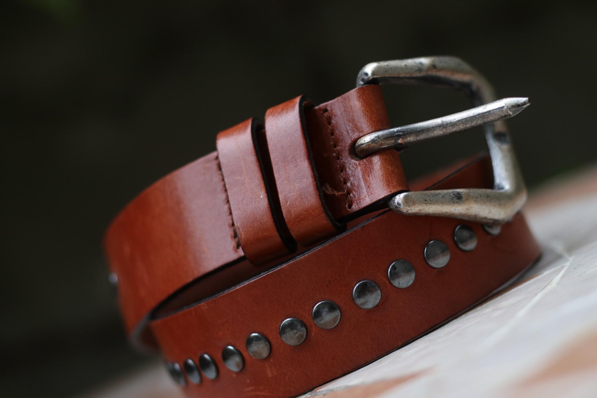A brown leather belt featuring a single row of metal studs along its length, curled on a patterned surface.