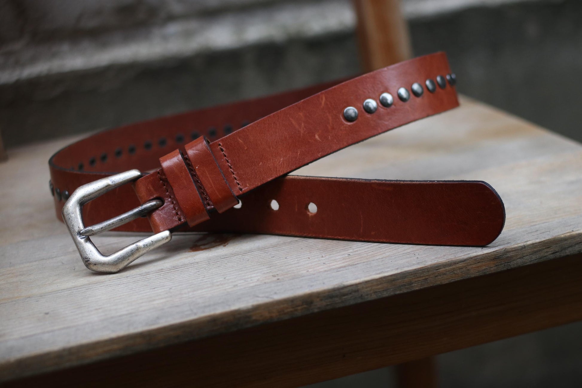 A vintage brown leather belt with a western flair, silver buckle, and decorative silver studs is presented on a textured wooden surface