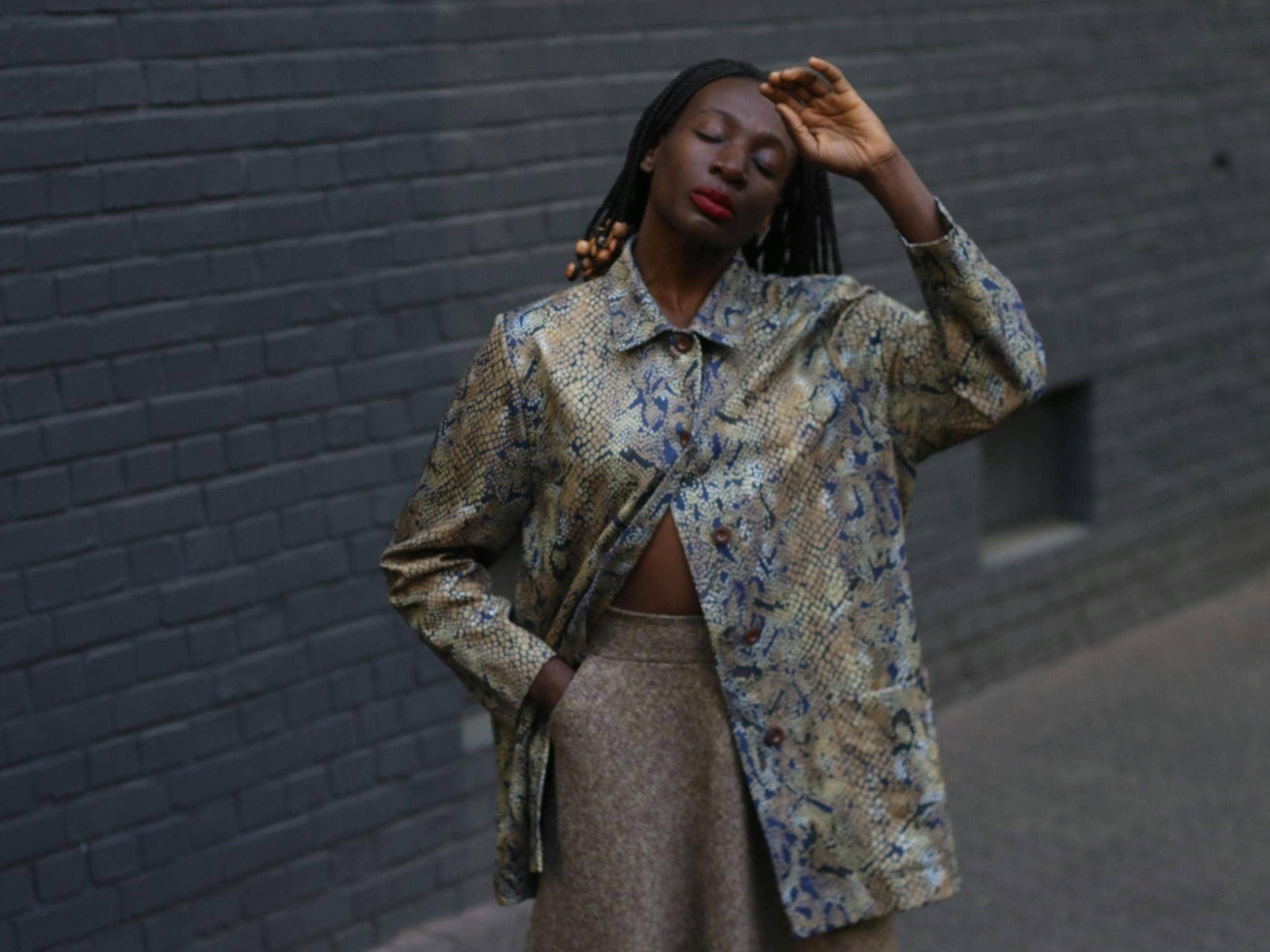 Female model with beaded braids  in a vintage 90s snakeskin duster jacket with a relaxed fit and oversized pockets photographed against  a grey brick wall to showcase the bold yet neutral layering piece perfect for standout street style