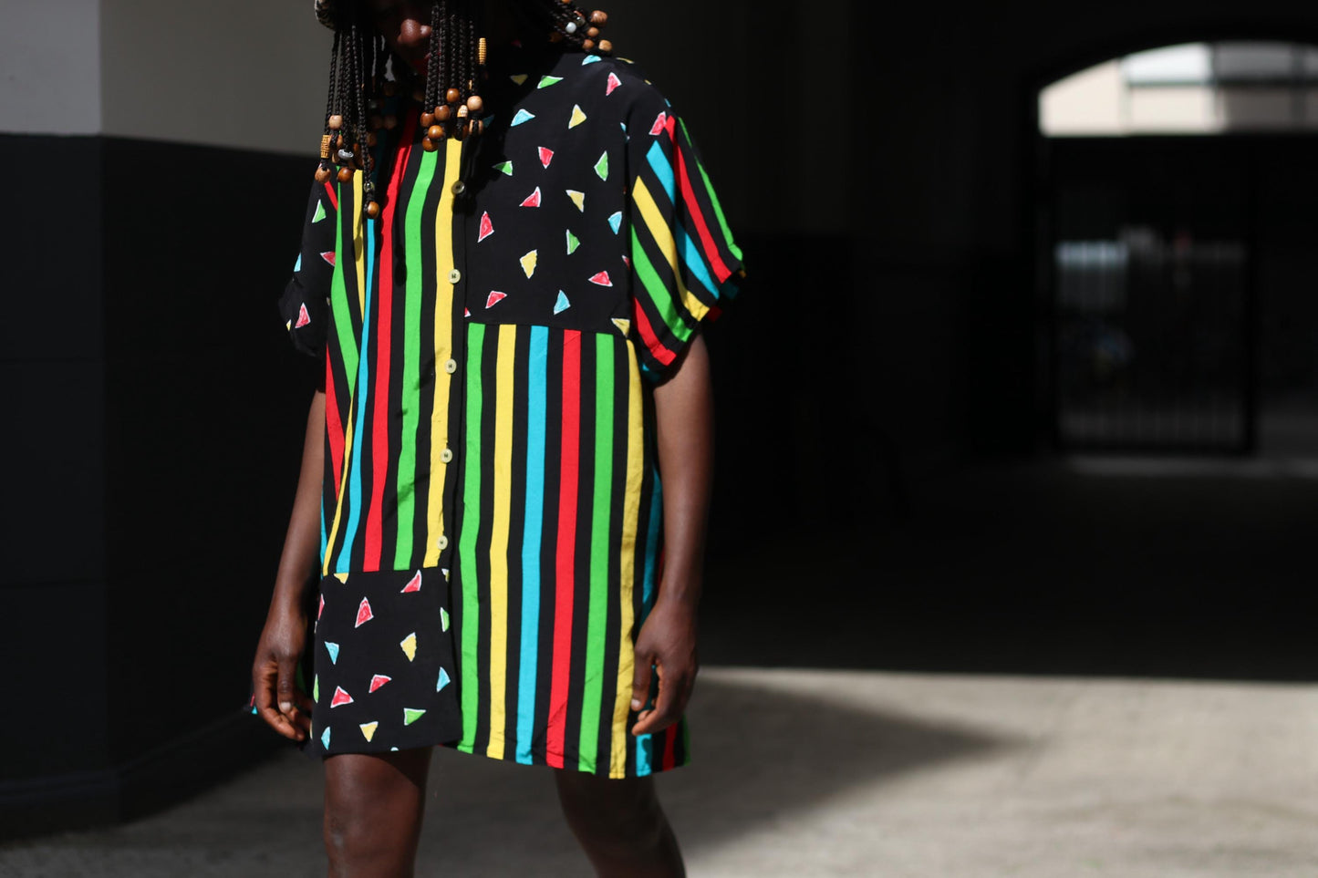 A close-up image of a female model in a colorful 90s oversized shirt worn as a dress with black mini leggings under. The retro button-up has  vertical stripes of red, yellow, green, and blue and features black panels with colorful triangles.  The image is shot in a sunlight alley