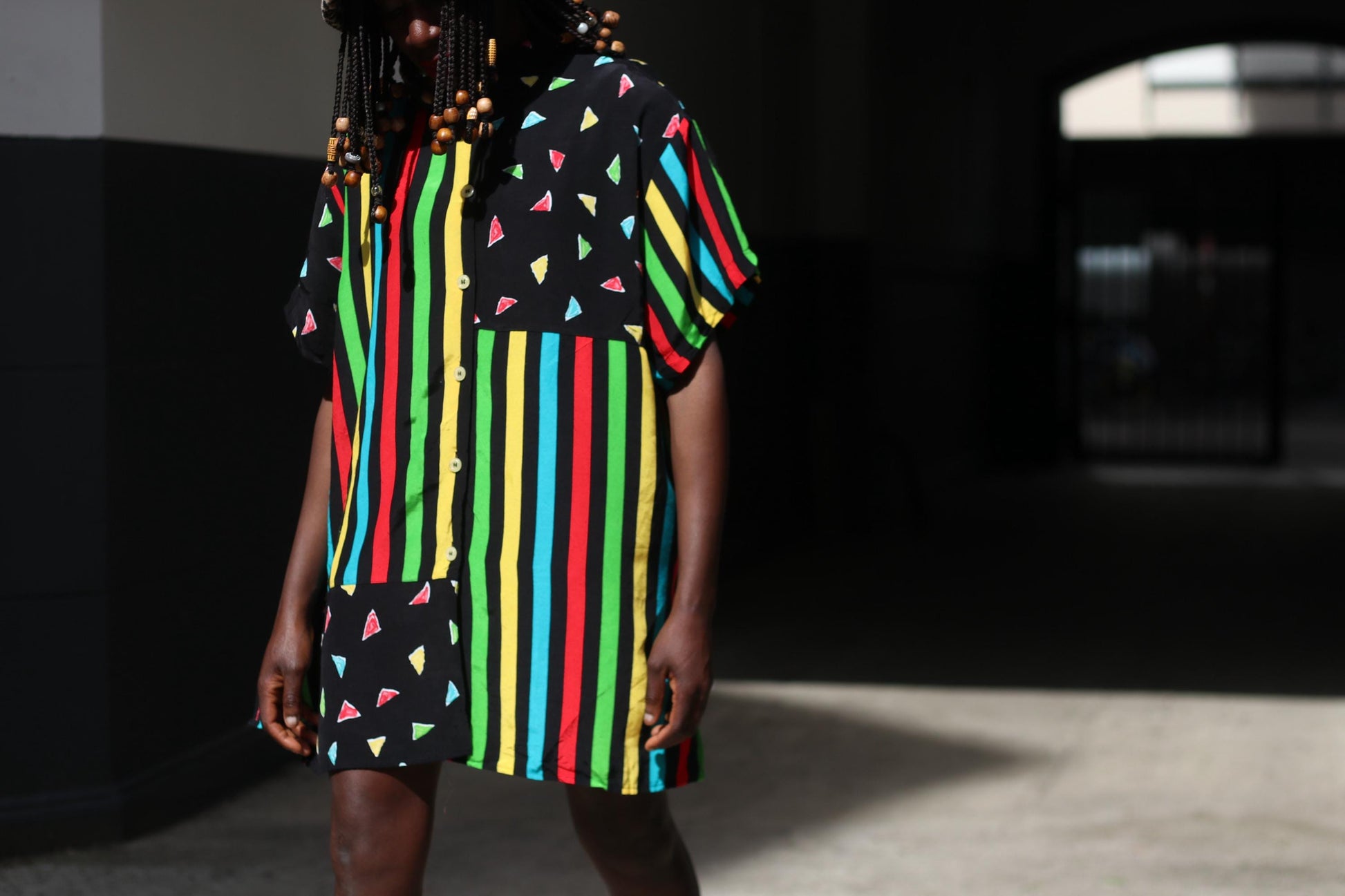 A close-up image of a female model in a colorful 90s oversized shirt worn as a dress with black mini leggings under. The retro button-up has  vertical stripes of red, yellow, green, and blue and features black panels with colorful triangles.  The image is shot in a sunlight alley