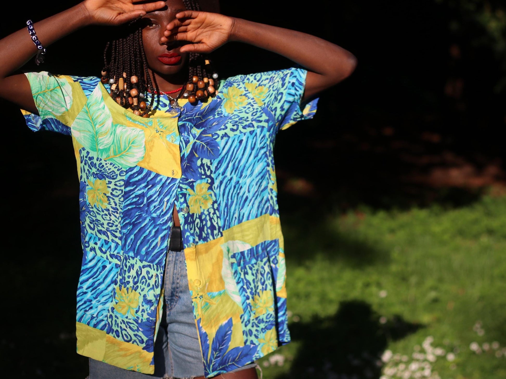Artistic image of a female model with beaded braids wearing a vintage Christine Laure Paris tropical print blouse in blue, yellow, and green, styled outdoors in sunlight.  Model has one hand covering her eye.