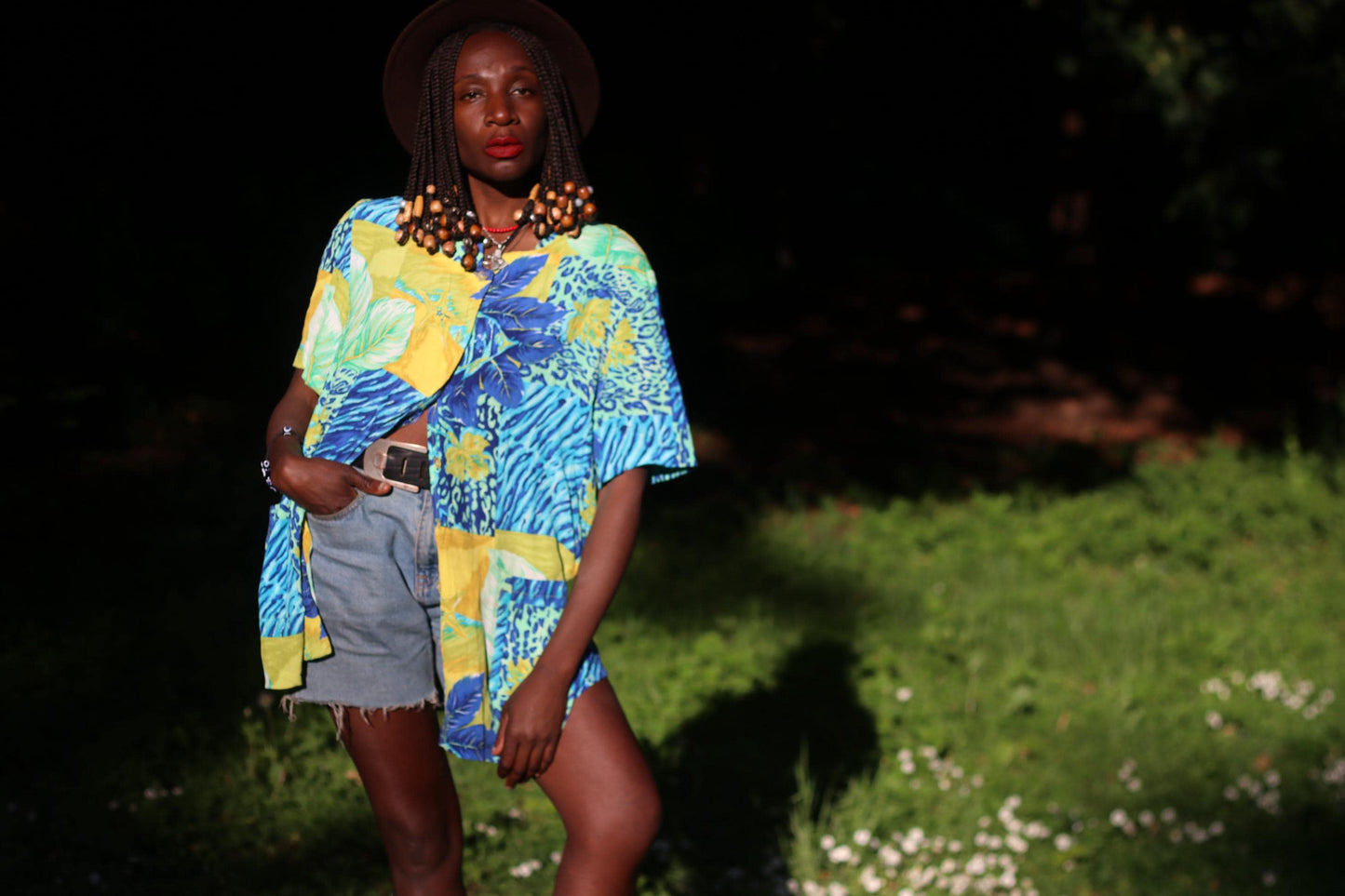Female model with beaded  braids wearing a french tropical shirt with vivid blue, green, and yellow prints, styled casually with blue  denim shorts in a green field with white daisies