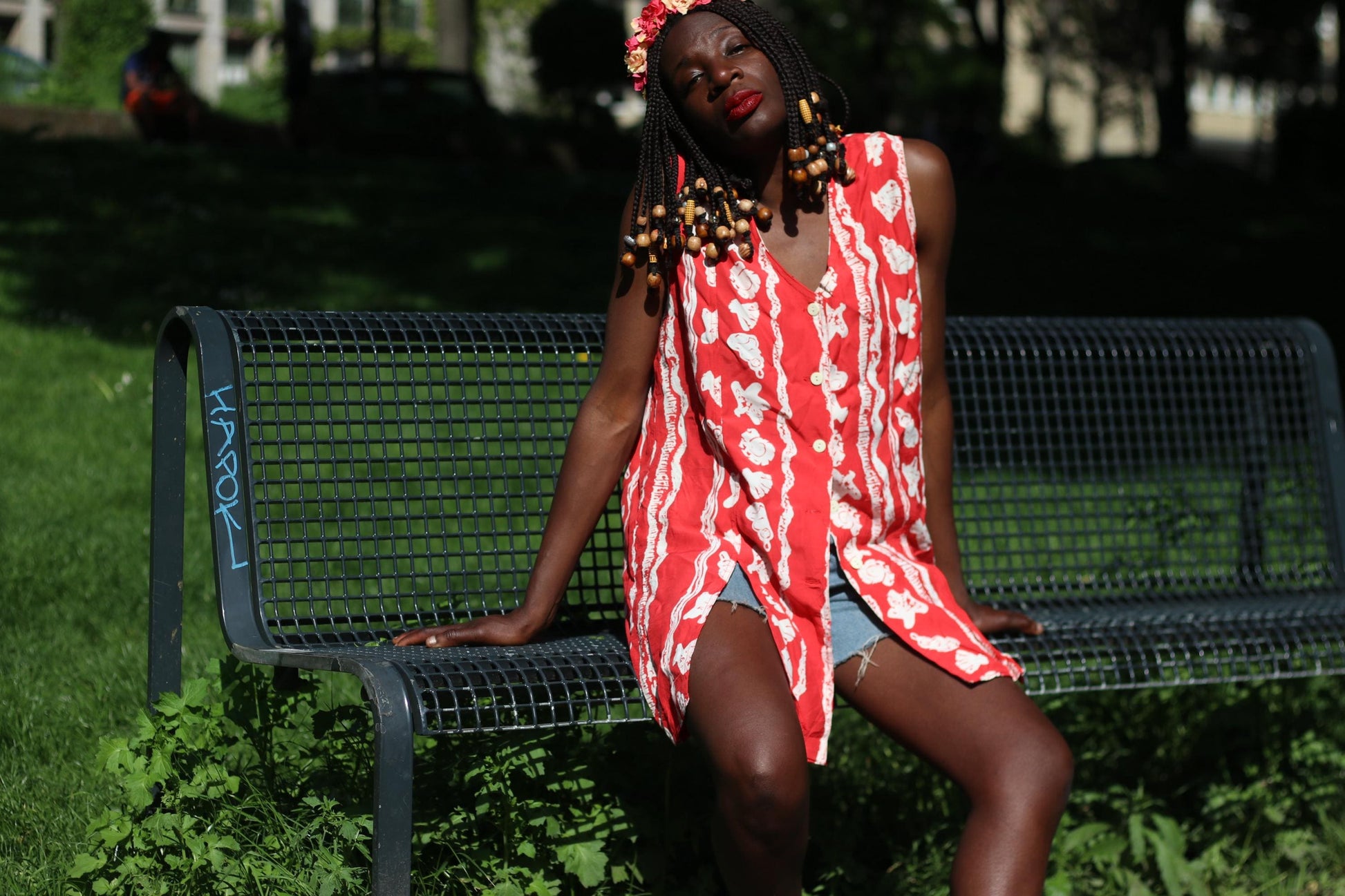 Front view shot of a person sitting on a green park bench, showcasing a vintage  red and white floral sleeveless top against a backdrop of green grass and trees. She wears blue denim shorts and a floral crown with the retro button-up