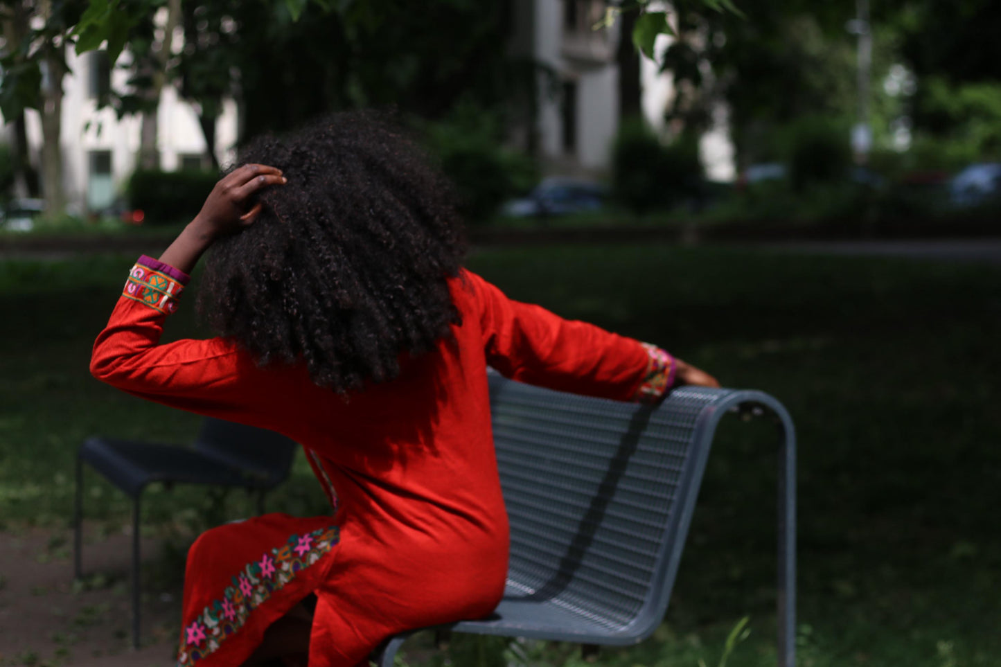 Back view of a woman wearing a vintage red embroidered boho maxi dress  with two side slits sitting on a park bench