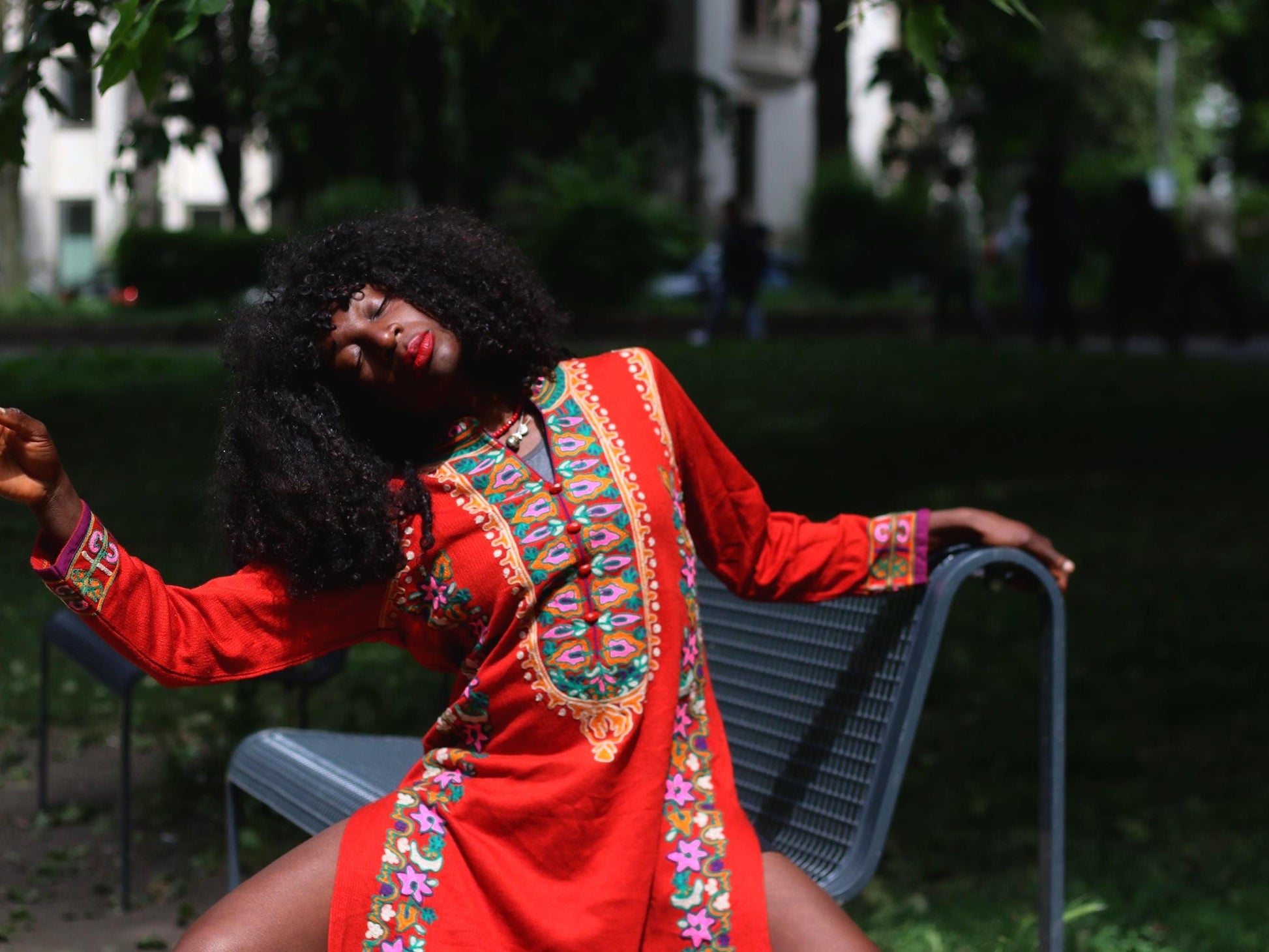 Woman with curly black hair wearing a vintage red embroidered boho maxi dress on a park bench.