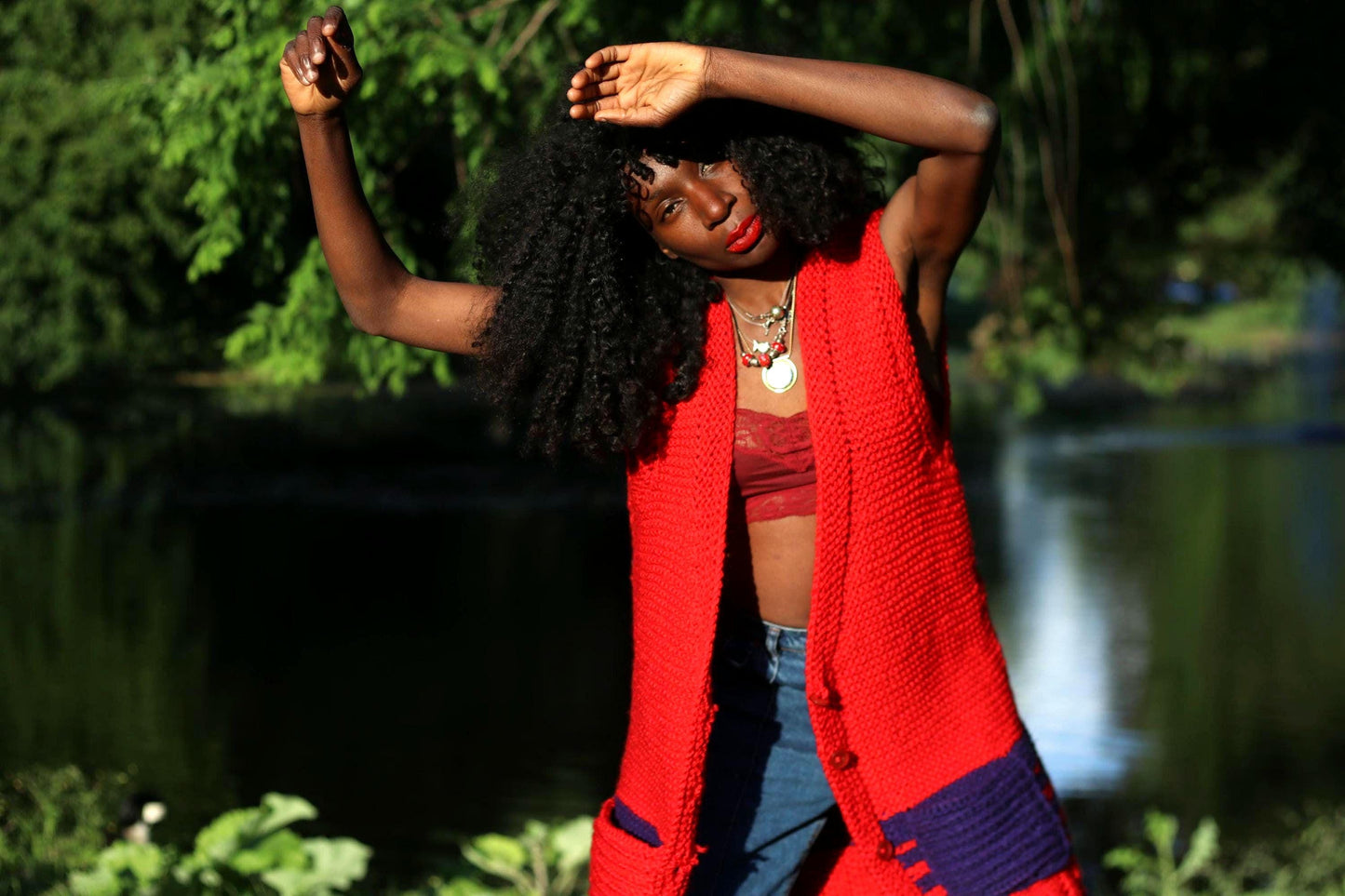 Female model is photographed outdoors in a park in front of a lake wearing a red vintage knitted vest with purple pockets over a cropped red lace-like top and styled with denim. The image gives off boho festival chic vibes