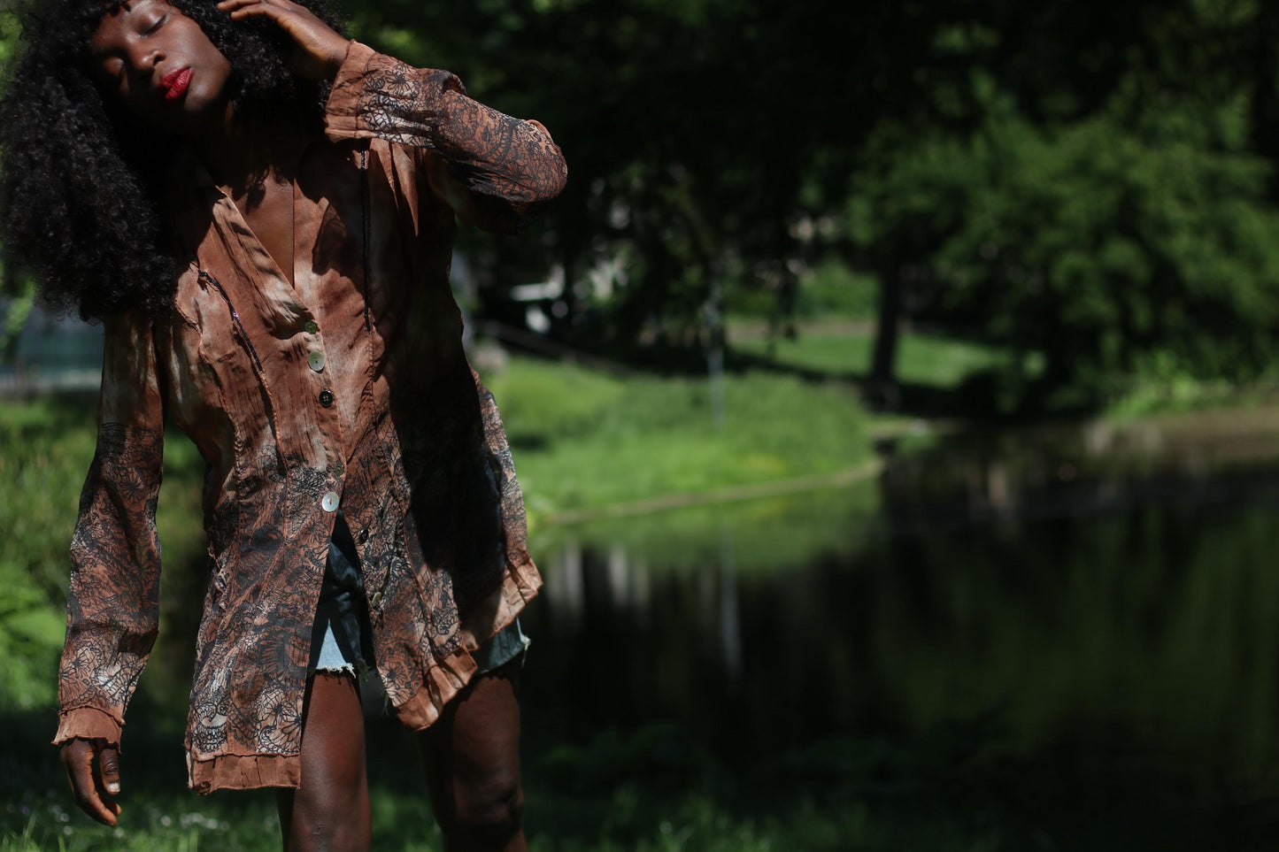 Female model with curly black hair wearing sheer brown BibA bohemian sheer blouse styled with denim shorts in nature setting outdoors with lake backdrop