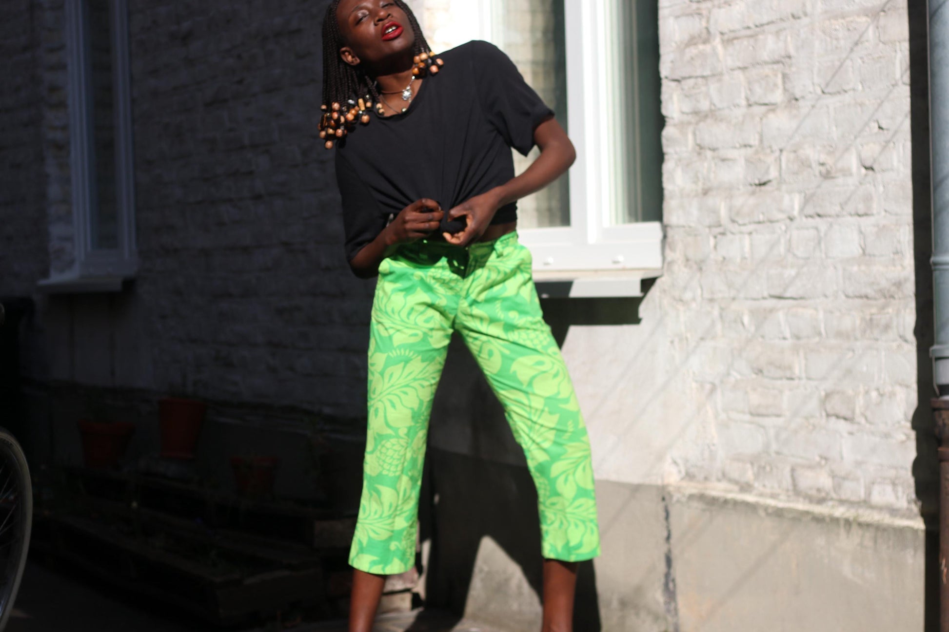 Happy model with beaded braids  wearing a  vibrant green floral capri pants by quiksilver standing outdoors by a white bricked wall.
