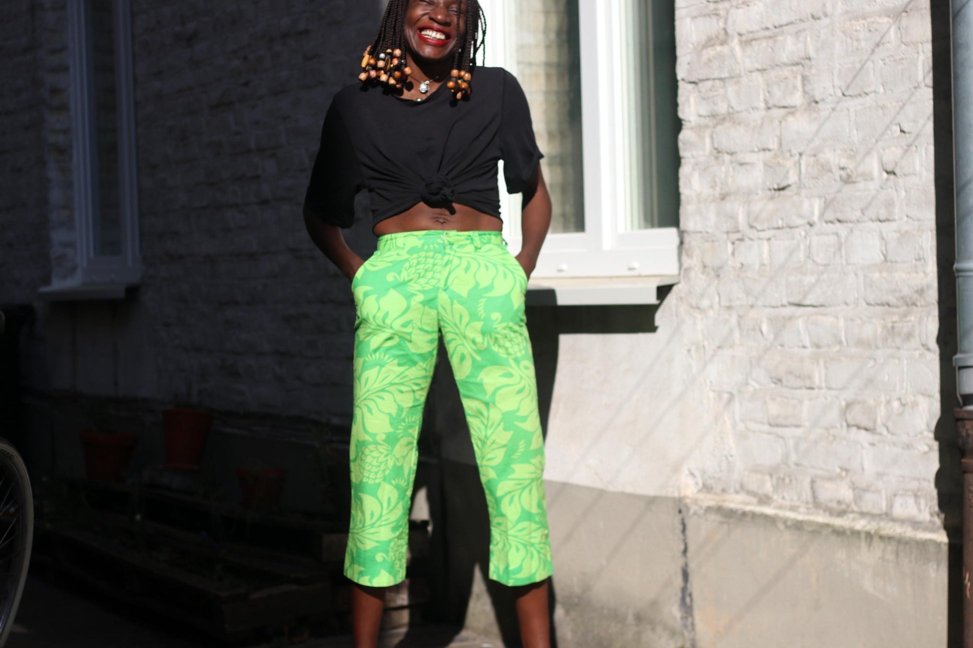 A female model smiling and standing, wearing vibrant green floral cropped pants and a black knotted t-shirt, against a light brick wall.